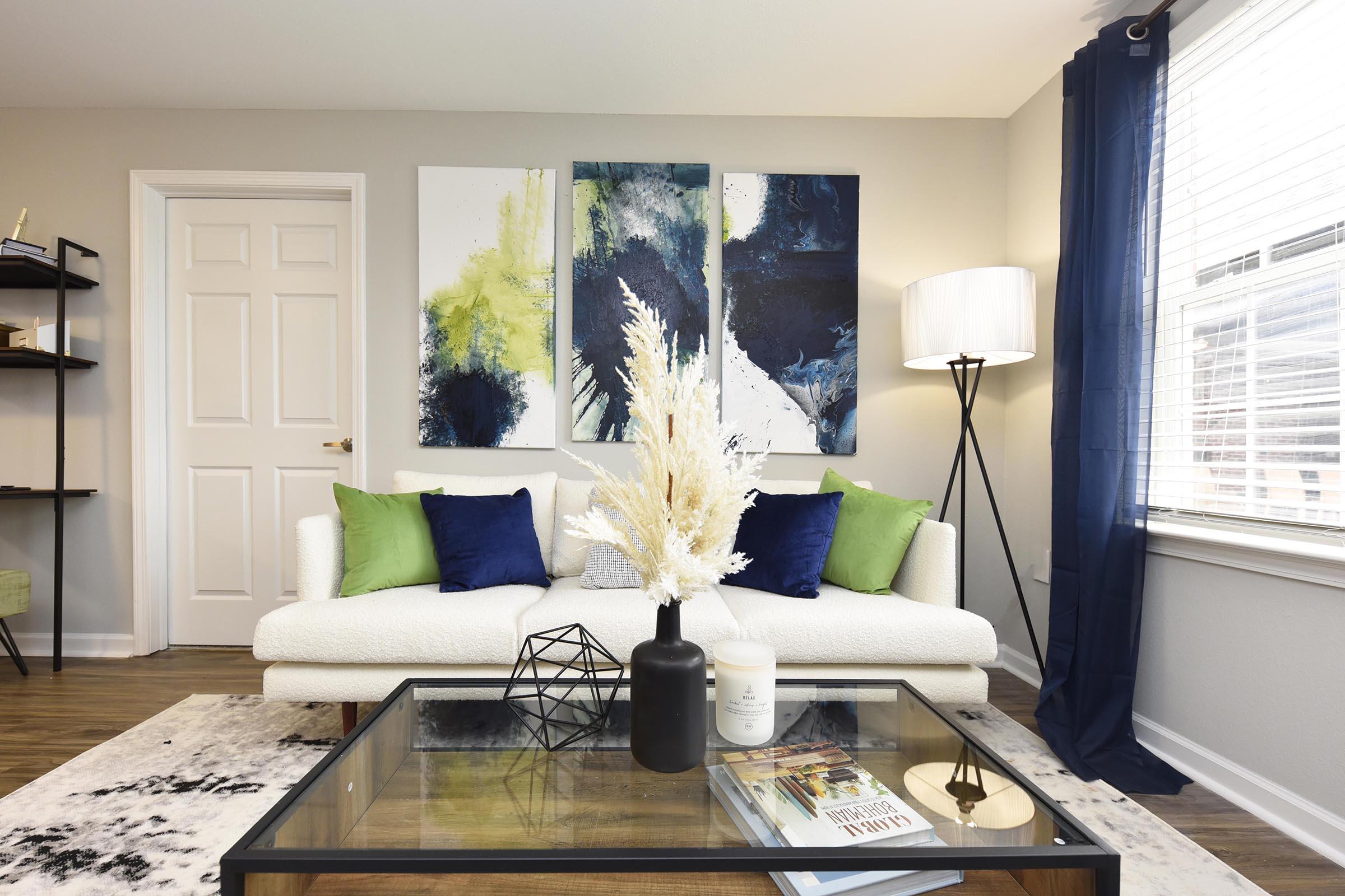 A modern living room featuring a white sofa with green and blue throw pillows, a glass coffee table with decorative items, and a vase of dried flowers. There are three abstract paintings on the wall, a stylish floor lamp, and a bookshelf in the background. Natural light filters through the window with blue curtains.