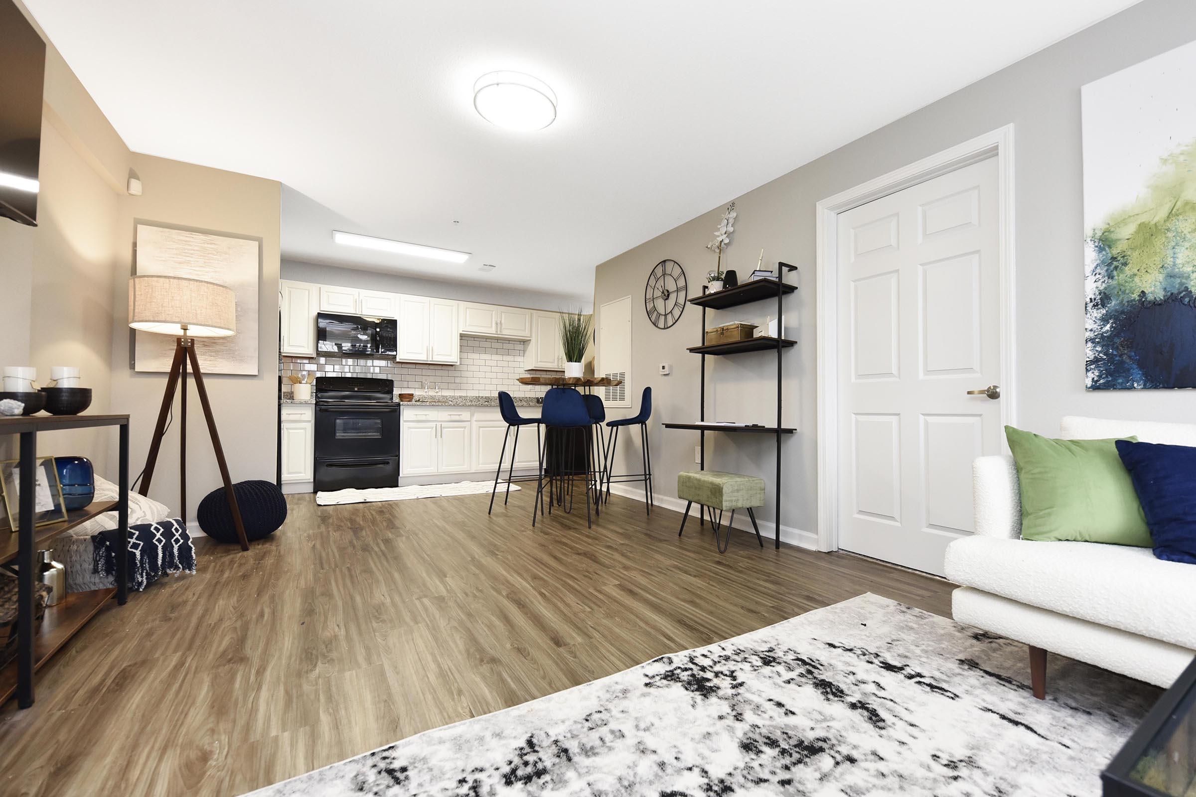 Modern living room and kitchen area with light wood flooring, featuring a sofa, small dining table with bar stools, a decorative shelf, and wall art. The kitchen is equipped with white cabinets and black appliances, while a large area rug adds comfort to the space.