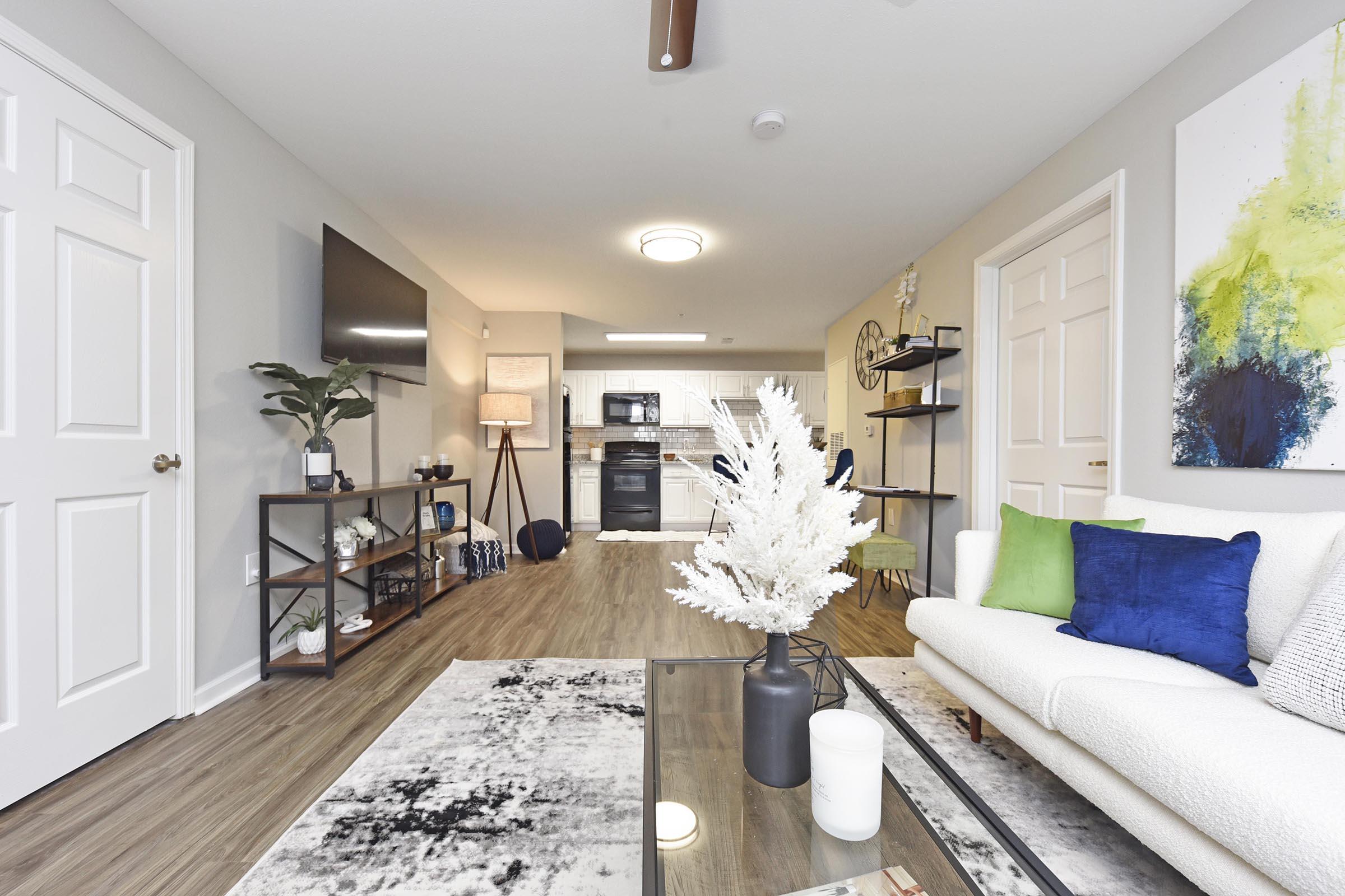 Modern living room featuring a plush sofa with colorful throw pillows, a glass coffee table, and a decorative vase. In the background, a kitchen is visible with dark appliances and white cabinetry. The space is bright and inviting with wood-like flooring and a large wall-mounted TV. Decorative plants and art pieces add a cozy touch.