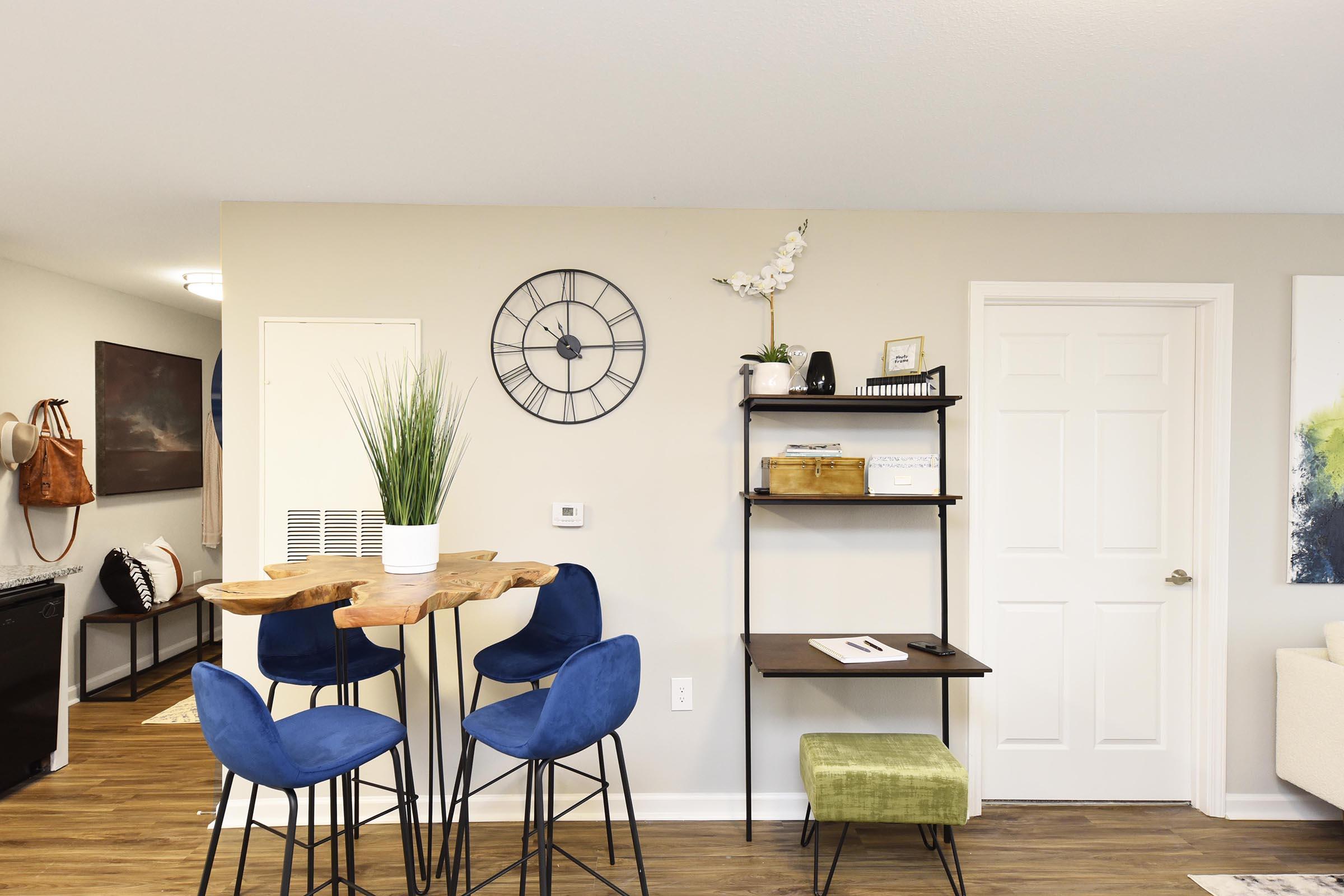A modern interior space featuring a dining area with four blue chairs around a wooden table. There's a clock on the wall, a small bookshelf with decorative items, and a plant. The room has light-colored walls and wooden flooring, creating a bright and inviting atmosphere.
