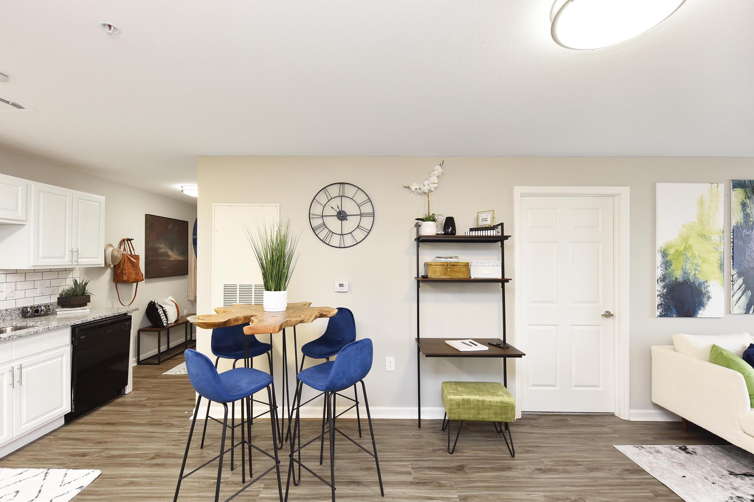 A modern kitchen and living area featuring a small dining table with blue chairs, a clock on the wall, and minimalistic shelving. The space is well-lit with natural light, showcasing a green plant and decorative artwork. A cozy couch is also visible, creating a stylish and inviting atmosphere.
