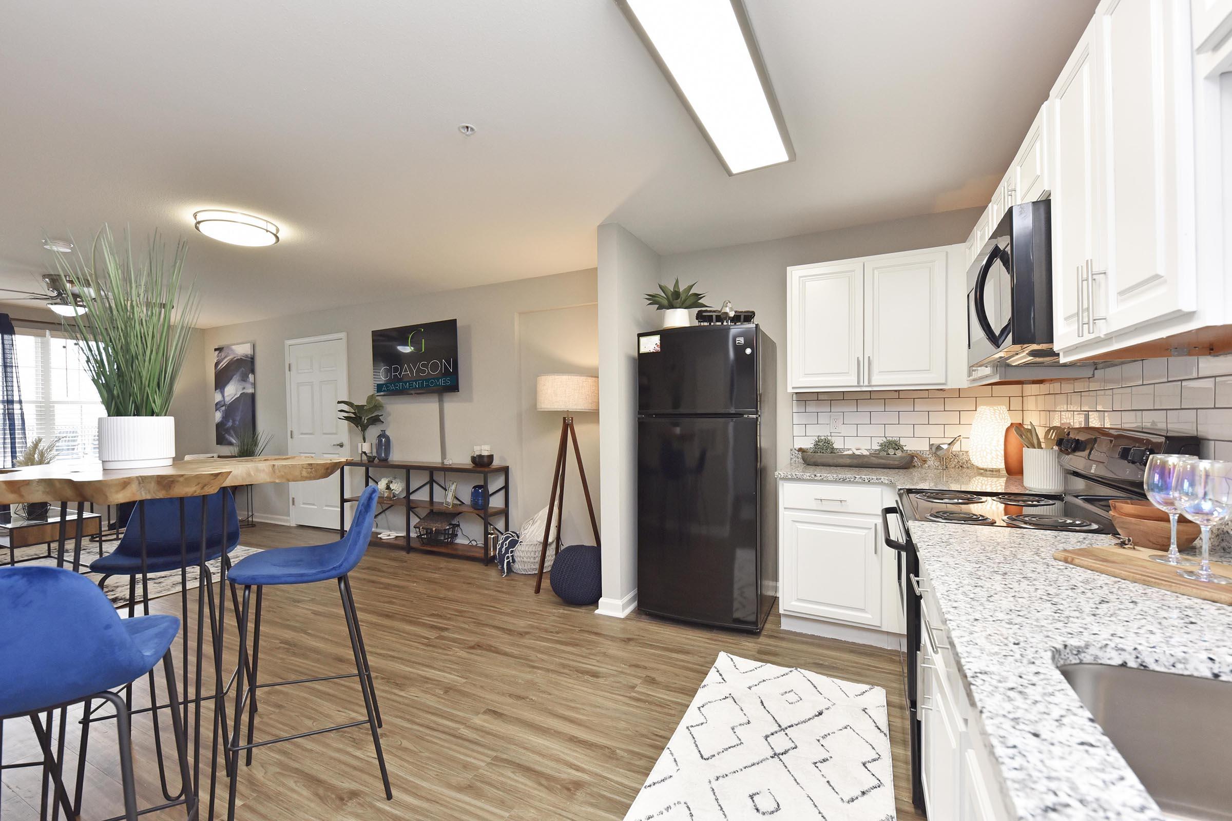 Modern kitchen featuring black appliances, white cabinetry, and granite countertops. A dining area with a wooden table and blue bar stools is visible. The space is well-lit with natural light and decorative elements like plants and artwork add a stylish touch.