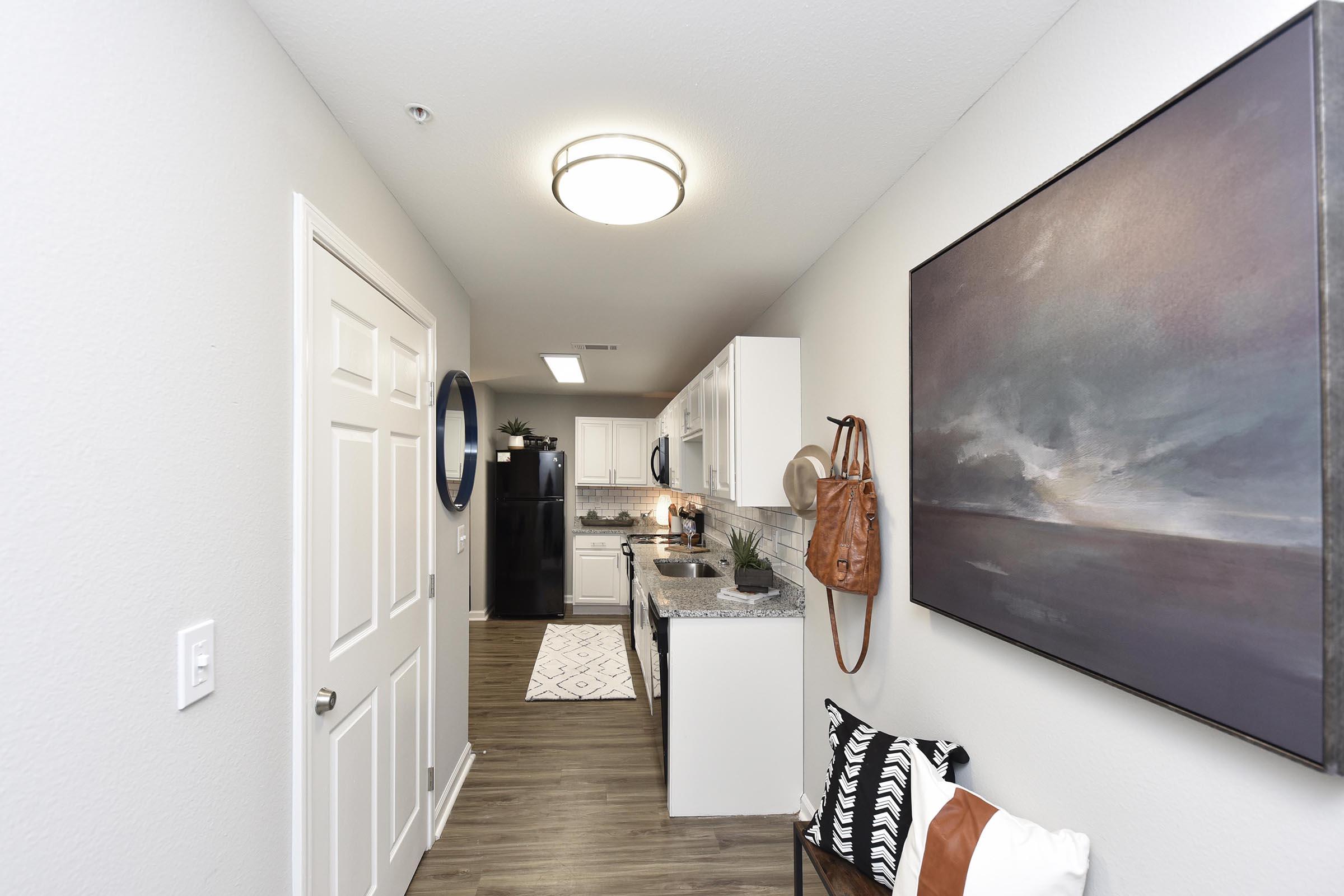 A modern kitchen featuring white cabinetry, a black refrigerator, and a granite countertop. The space includes a decorative painting on the wall and a stylish chair with patterned cushions. Natural light illuminates the area, creating a welcoming atmosphere.