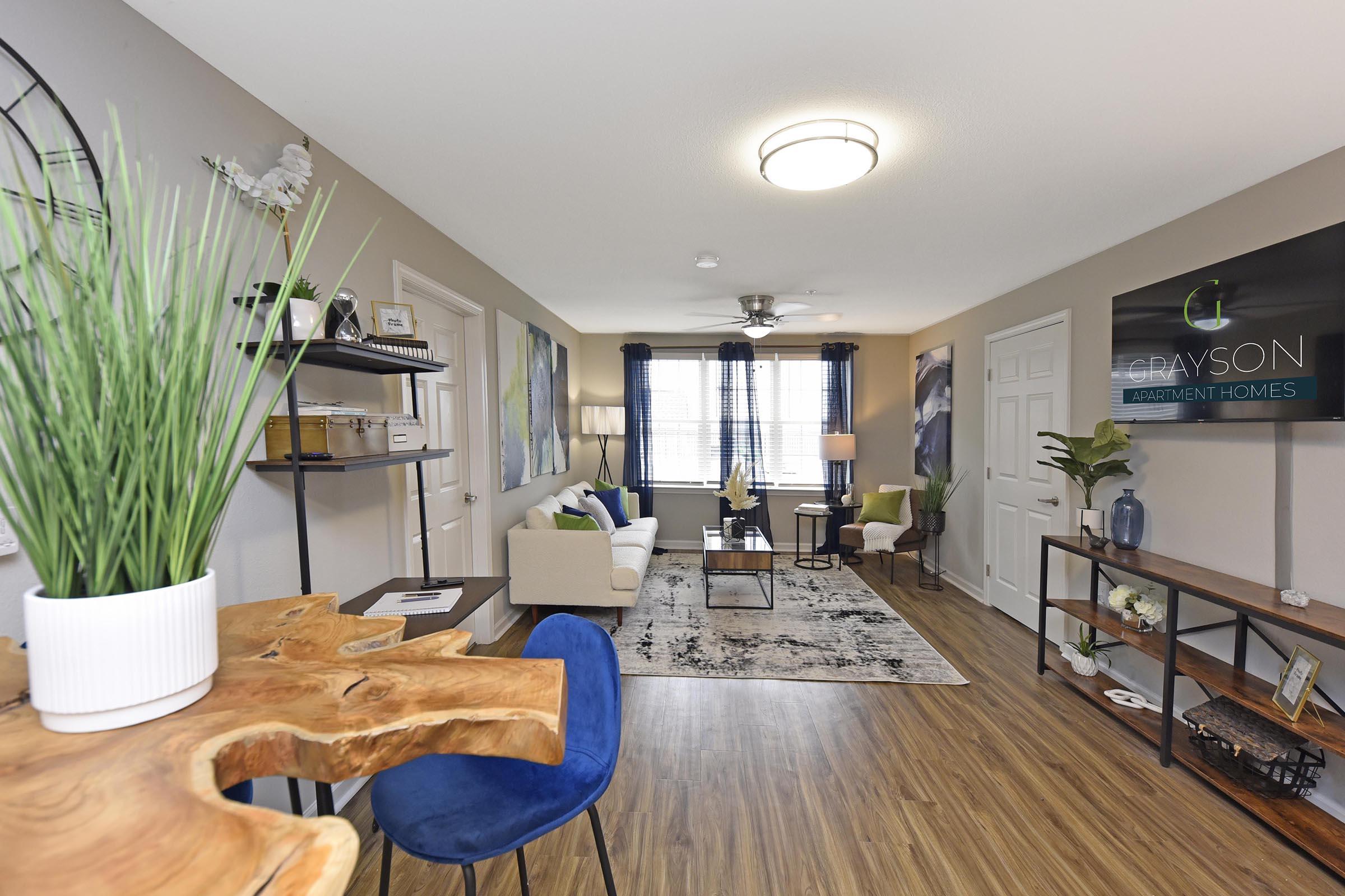 A modern living room in an apartment featuring a beige sofa, decorative cushions, a patterned area rug, and a wooden table. Windows with blue curtains provide natural light, while a potted plant adds a touch of greenery. A wall sign displays "Grayson Apartment Homes." 