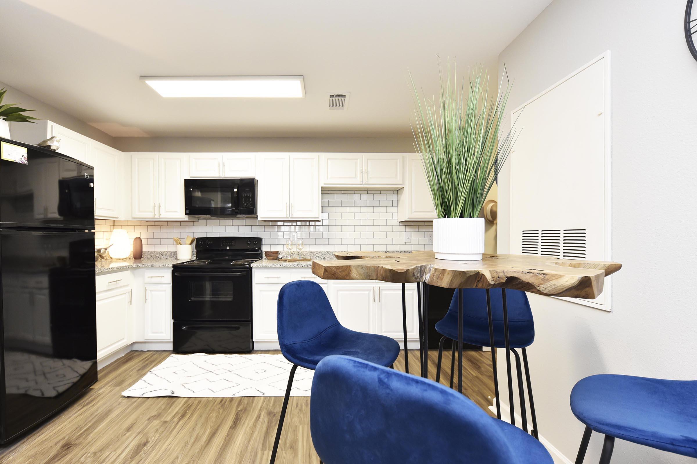 A modern kitchen featuring white cabinets, a black refrigerator, and an oven. A wooden table with a unique design and blue chairs is set nearby. The kitchen has a light-colored countertop, a white backsplash, and a potted plant for decor, creating a stylish and inviting atmosphere.