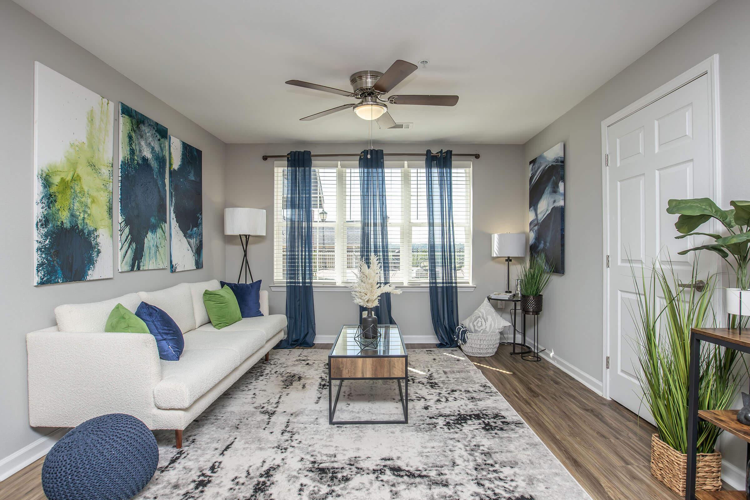 A modern living room featuring a light-colored sofa with green and blue cushions, a glass coffee table, and a decorative rug. Large windows with blue curtains allow natural light, and greenery accents add a fresh touch. Artwork in blue and green tones adorns the walls, creating a stylish, relaxed atmosphere.