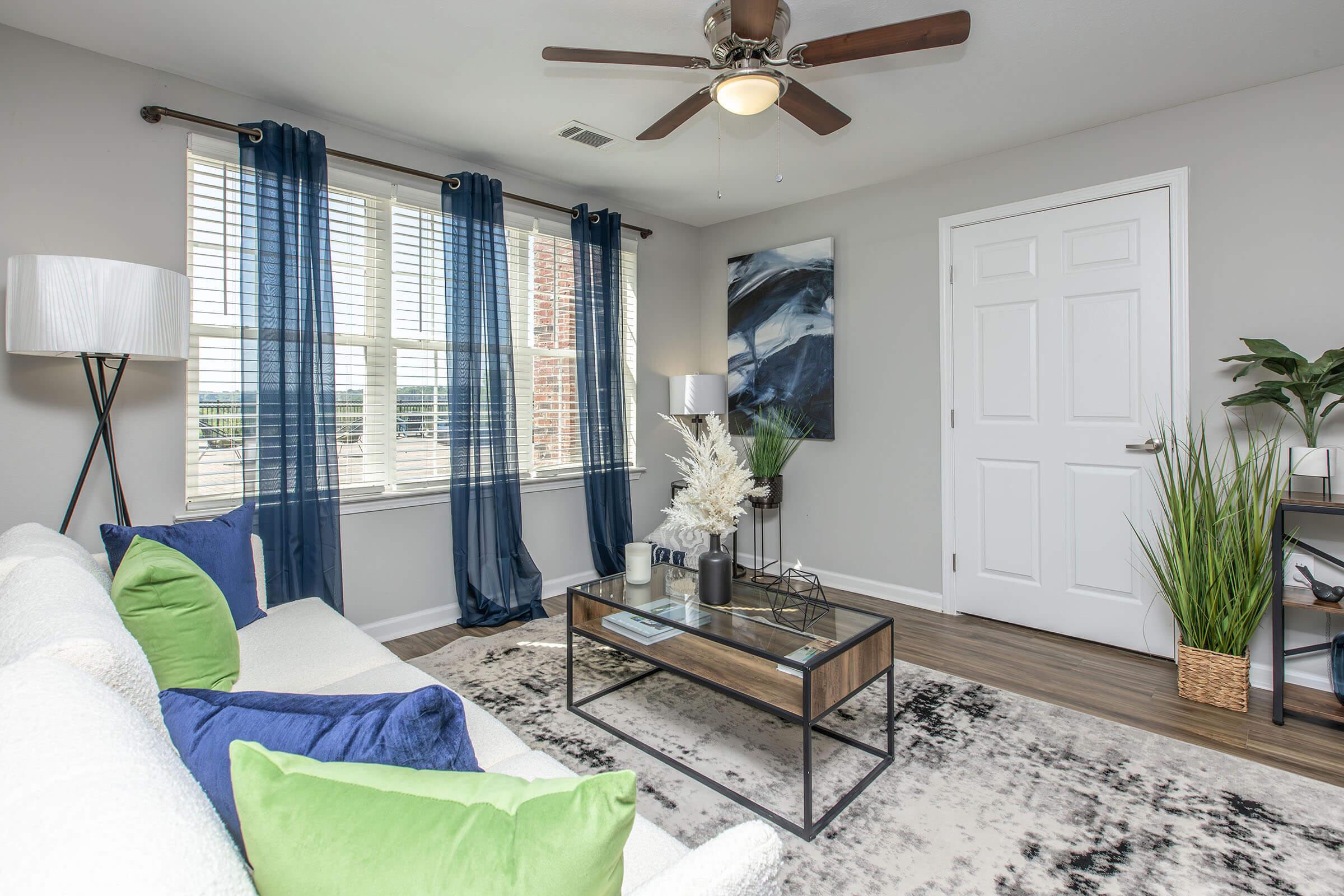 A modern living room featuring a white couch with blue and green pillows, a glass coffee table with decorative items, and a large window with blue curtains. There's a potted plant in the corner and a piece of abstract wall art. The space is well-lit and cozy, with wood-like flooring.