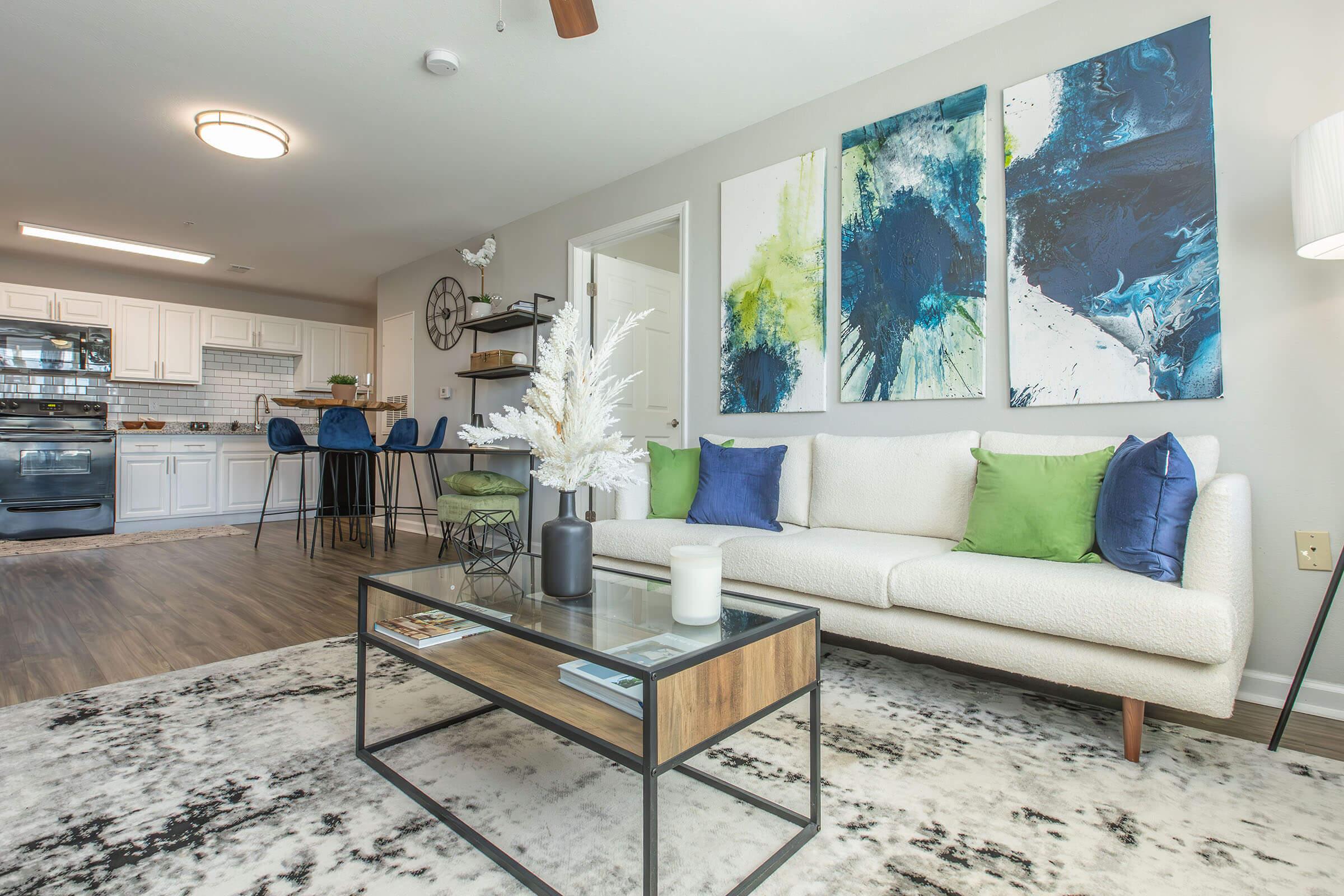 A modern living room featuring a white sofa with blue and green accent pillows. A glass coffee table sits on a patterned rug, with a decorative vase on top. Abstract art pieces in blue, green, and yellow adorn the wall. In the background, a kitchen area with white cabinets and bar stools is visible.