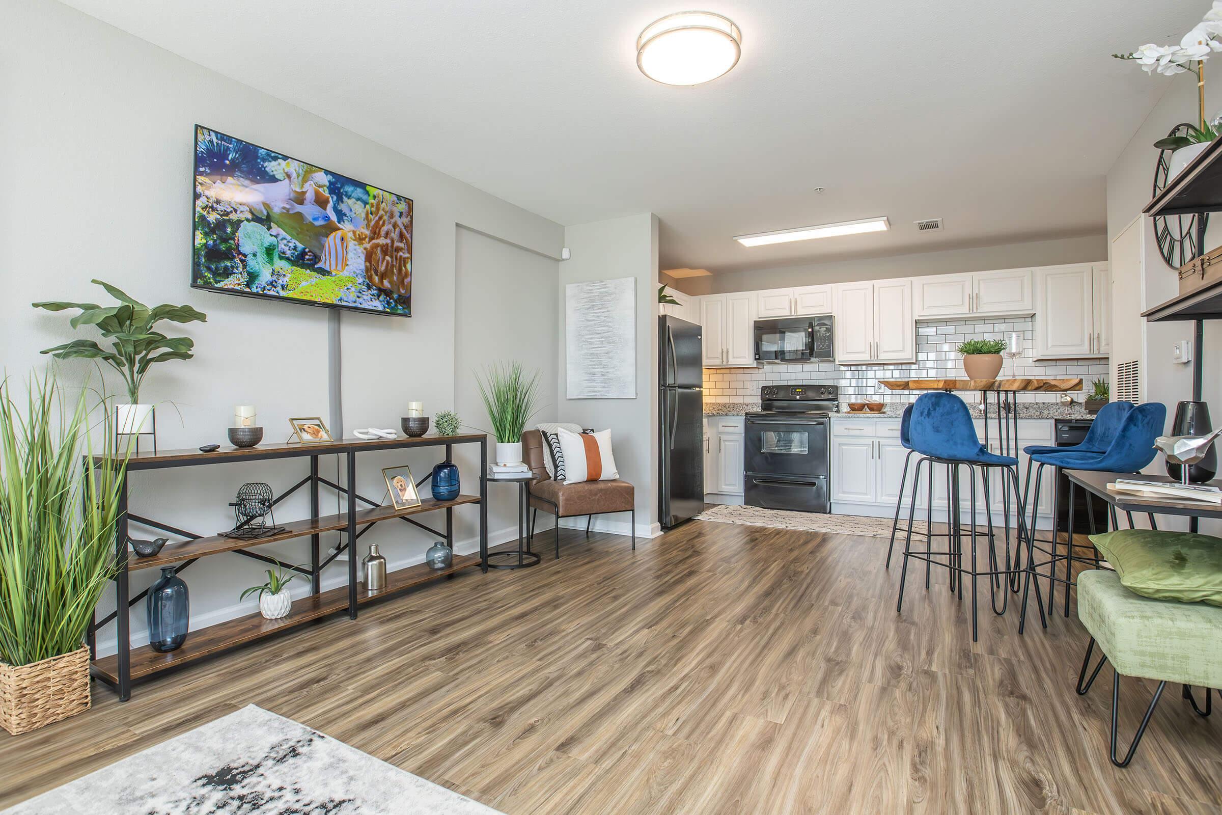 A modern living space featuring a cozy kitchen area with white cabinets and black appliances. There is a flat-screen TV mounted on the wall displaying nature scenery, a stylish shelf with decorative items, and bar stools at a counter. Lush plants add a touch of greenery, enhancing the inviting atmosphere.