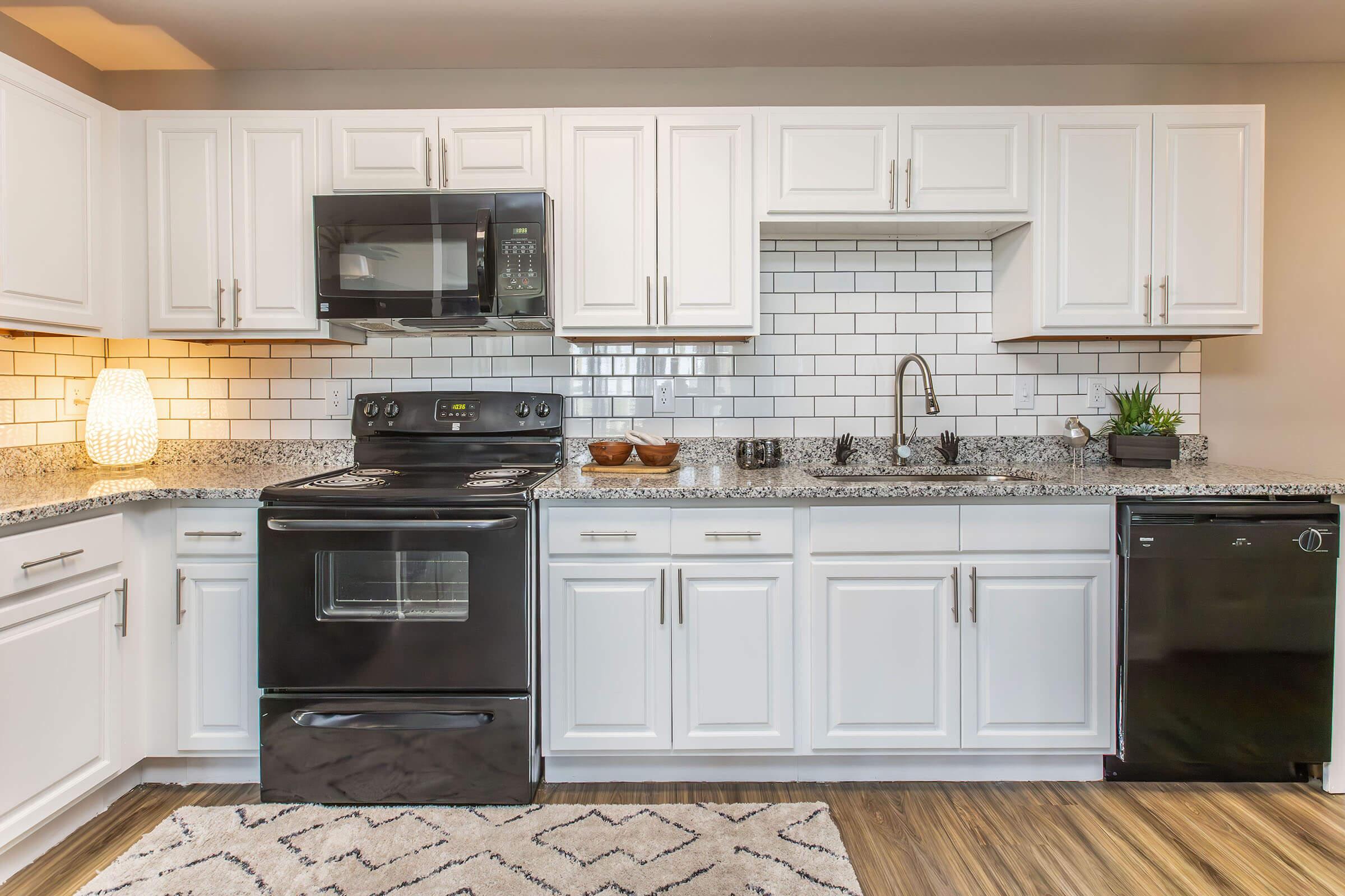 Modern kitchen with white cabinets, a black stove and microwave, a sleek sink, and a black dishwasher. The countertop features a gray granite pattern, paired with a white tile backsplash. A decorative lamp and a small plant add a touch of warmth to the space, which has a neutral color scheme and wood-like flooring.