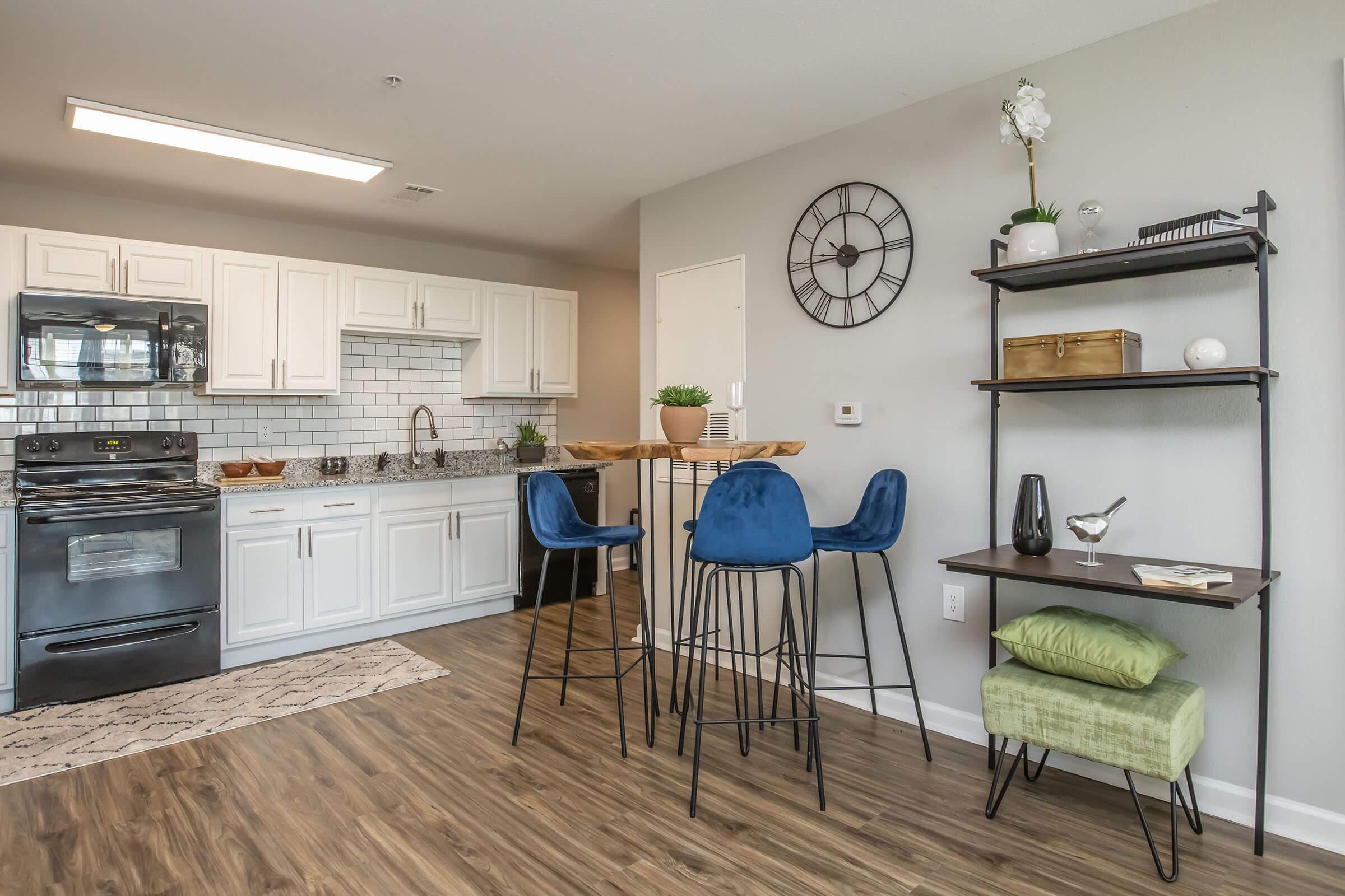 Modern kitchen and dining area featuring white cabinetry, a black stove, and a stylish clock on the wall. A small dining table with blue bar stools is set against a backdrop of wood flooring, complemented by decorative plants and a shelf displaying various items. Bright and inviting ambiance.