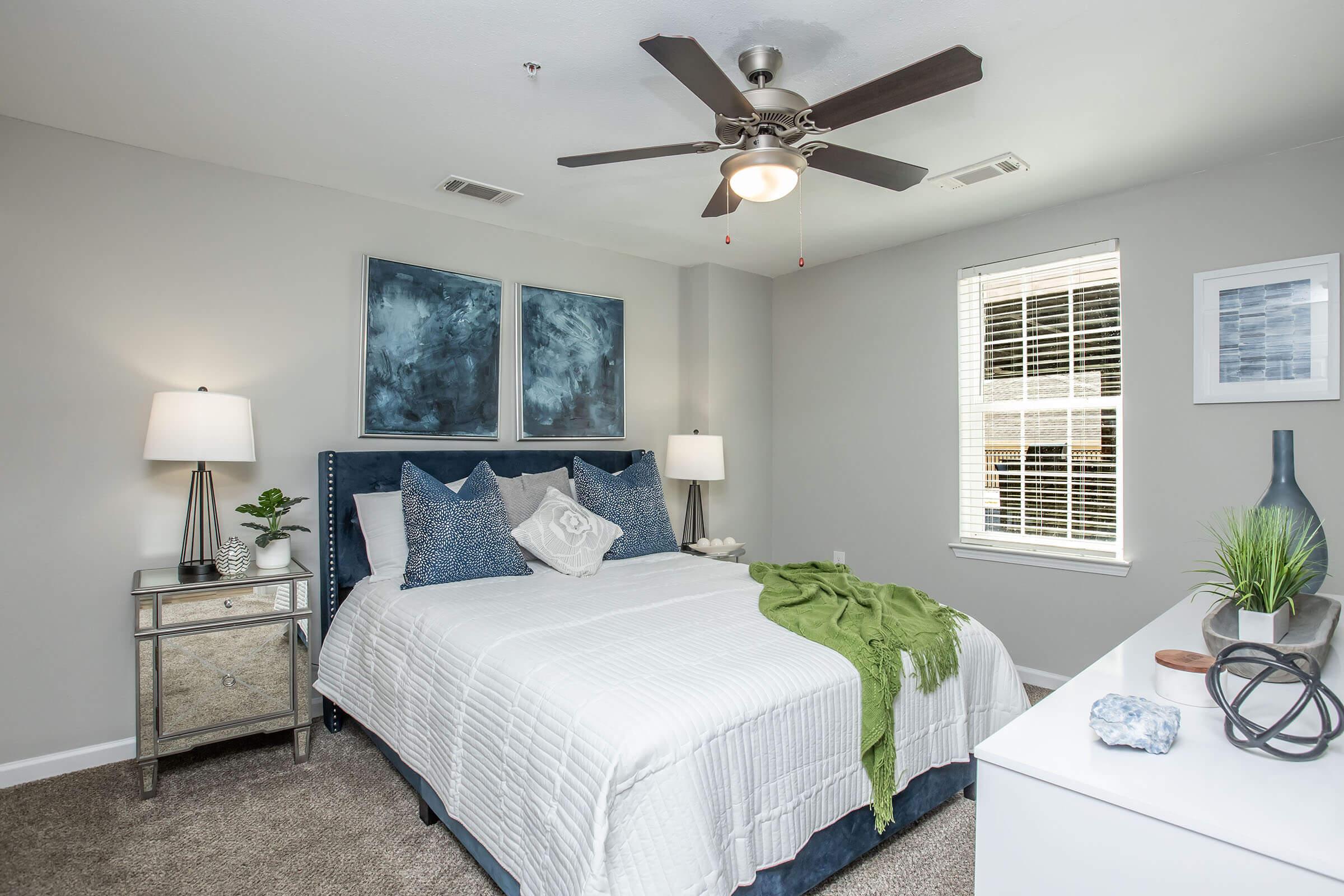 A modern bedroom featuring a queen-sized bed with decorative pillows and a green throw blanket. Two bedside lamps illuminate the space. The walls are painted light gray, and the room includes abstract artwork and a small plant on a nightstand. A ceiling fan and large window complete the cozy atmosphere.