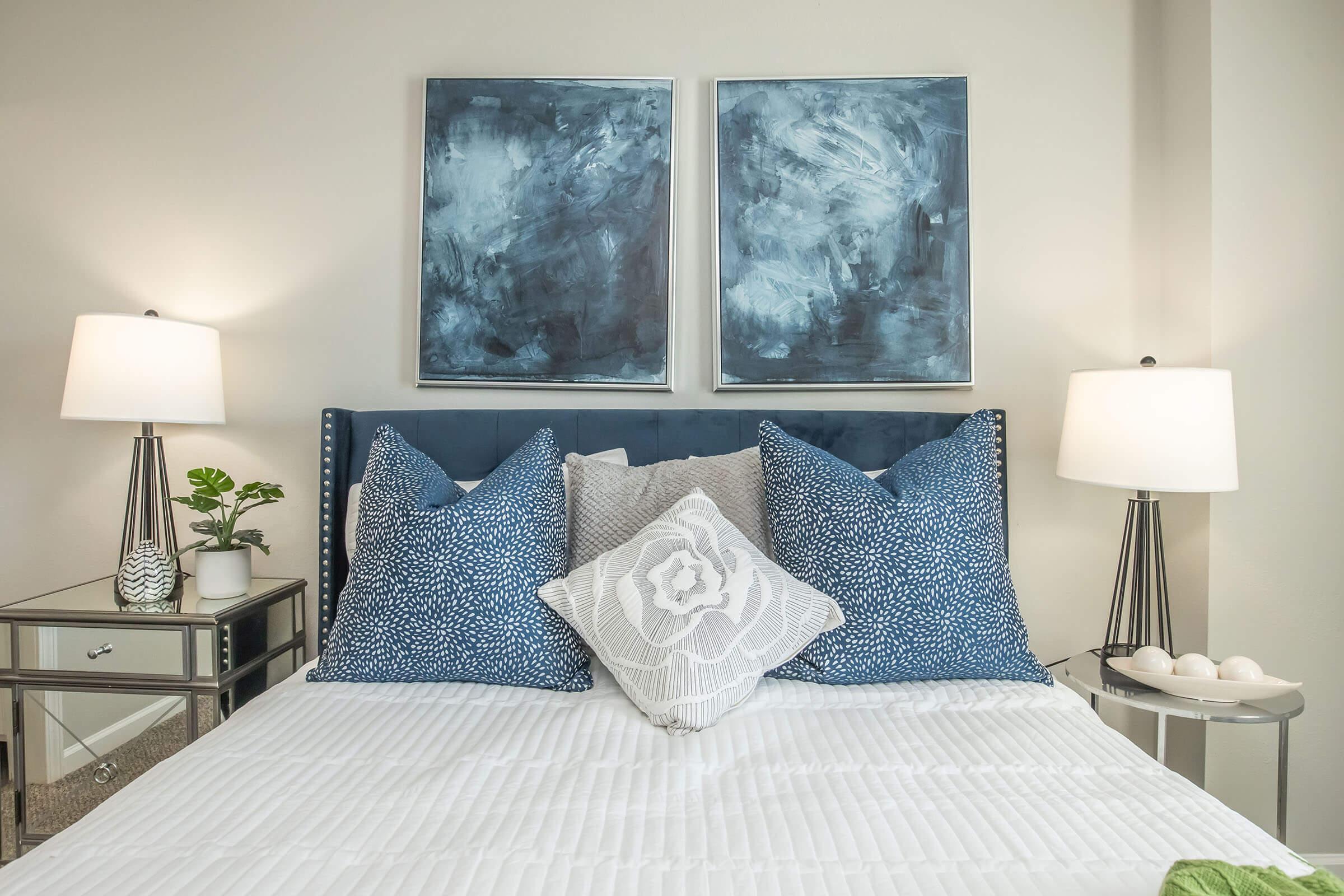 A modern bedroom featuring a neatly made bed with white bedding and decorative pillows in shades of blue. Two lamps on either side of the bed provide warm lighting. Above the bed are two abstract blue wall art pieces. A small potted plant and decorative items are on a mirrored nightstand.