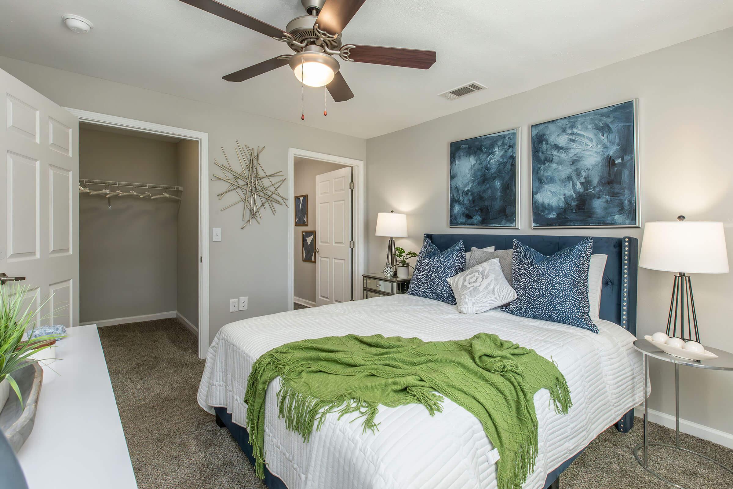 A cozy bedroom featuring a large bed with white bedding and a green throw, two modern lamps on either side, blue abstract artwork on the walls, and a ceiling fan. A closet is visible in the background, and there’s a decorative starburst mirror above a small dresser. The flooring is carpeted in neutral tones.