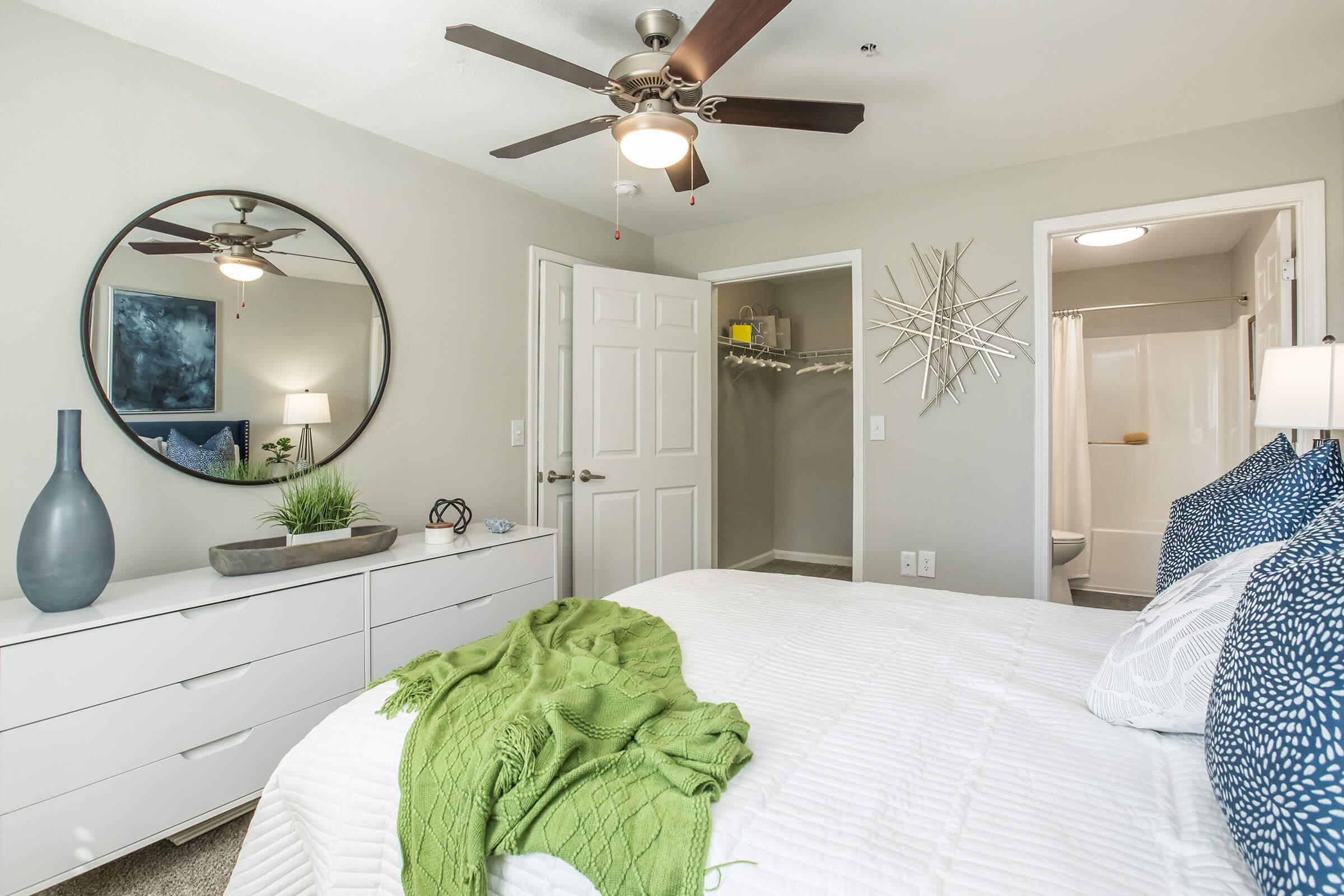 A modern bedroom featuring a white bed with a green throw blanket, a round mirror, and a ceiling fan. There is a decorative vase, a dresser, and a closet visible. The walls are painted in a light color, and a bathroom is seen through an open doorway.