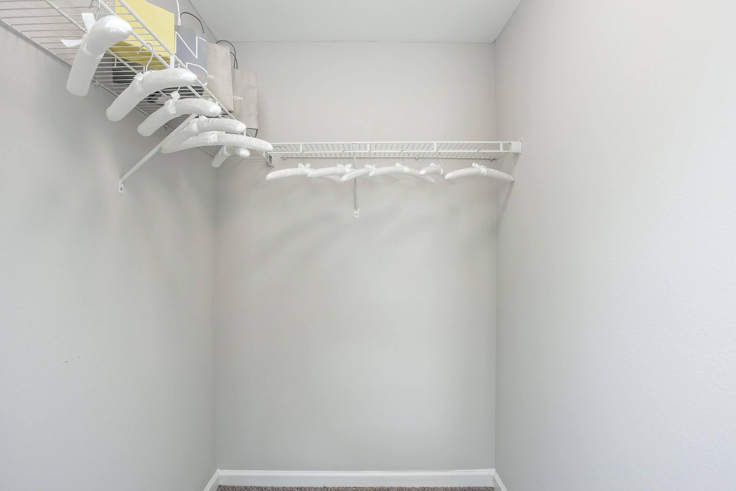 A clean, empty closet featuring a white wire shelf with several hanger hooks. The walls are painted a light color, and the floor is covered with carpet. The shelf holds a few items, including a small box, while most of the space remains clear, suggesting potential for organization or storage.