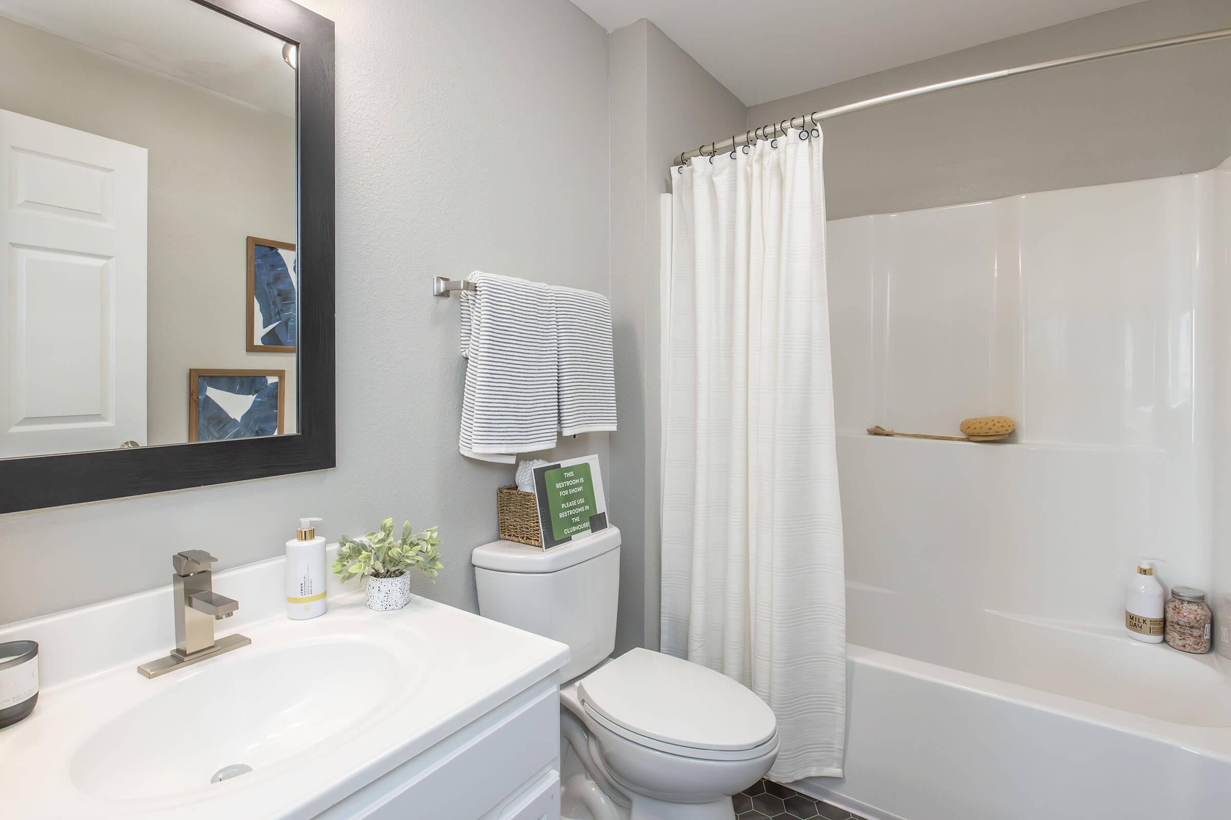 A modern bathroom featuring a white sink with a faucet, a white toilet, and a bathtub with a shower curtain. The walls are painted gray, and there's a mirror above the sink. A towel rack holds a neatly folded towel, and decorative items, including a small plant and framed pictures, are visible on the countertop.