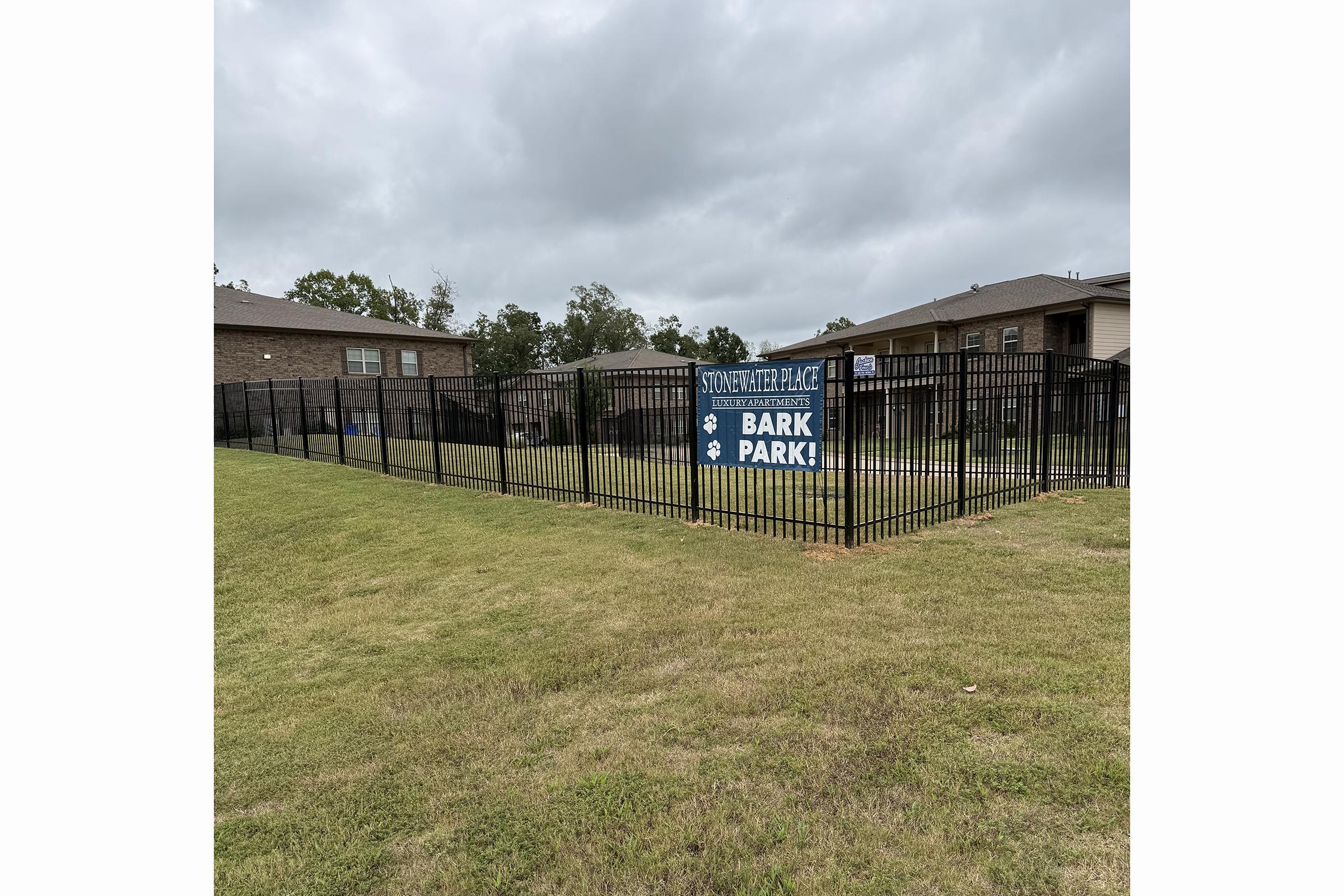 A fenced dog park featuring a sign that reads "Stone Ridge Place Bark Park." The park is surrounded by grass and a cloudy sky, with nearby residential buildings visible in the background.