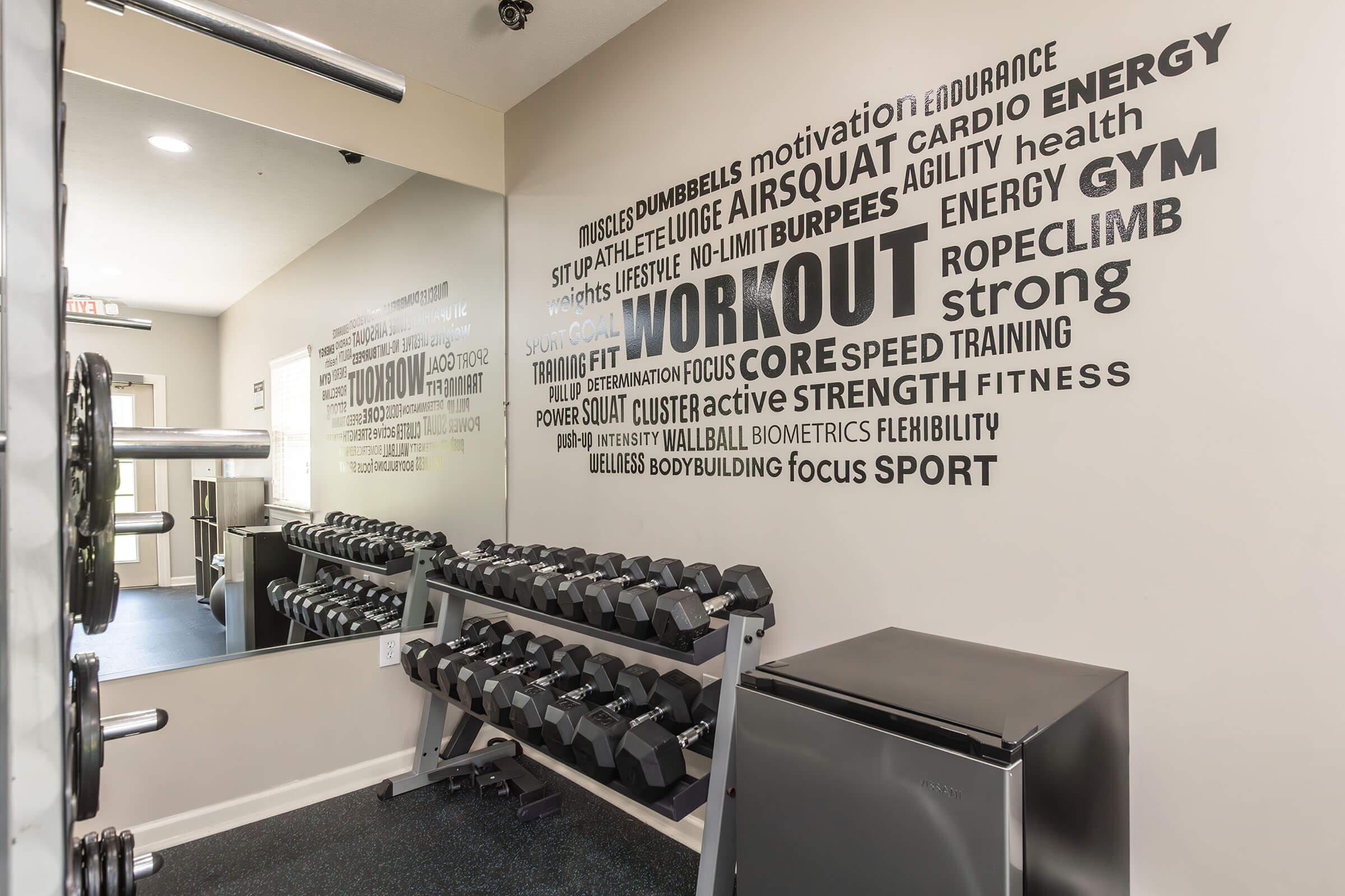 A fitness area featuring a rack of dumbbells against a wall decorated with various motivational fitness words and phrases like "WORKOUT," "STRENGTH," "CARDIO," and "ENDURANCE." A mirror reflects the space, emphasizing a focus on training and exercise.