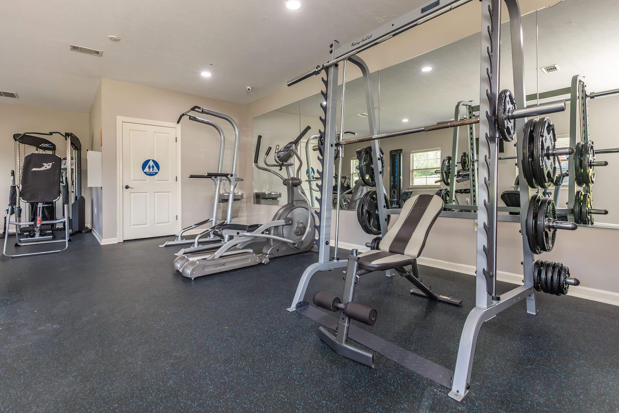 A well-equipped gym interior featuring a weightlifting bench, barbell with weights, an elliptical machine, and a mirror reflecting the space. The floor is dark and the walls are light, creating a bright and open atmosphere. A door in the background leads to another area of the facility.