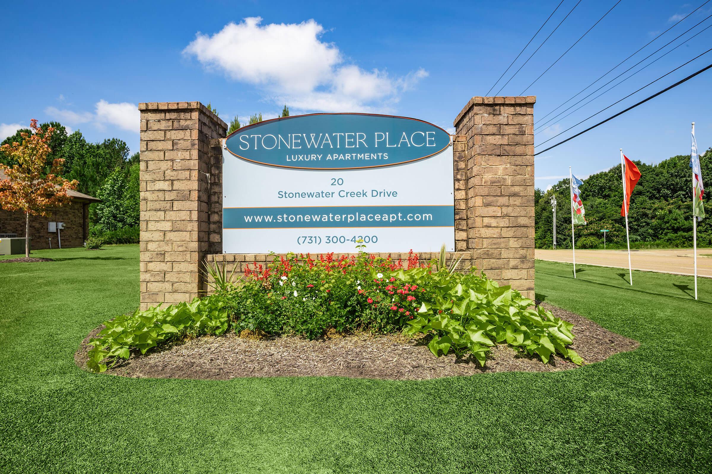 Sign for Stonewater Place Luxury Apartments, featuring the address "20 Stonewater Creek Drive" and a contact number "(731) 300-4200." The sign is set against a blue sky with some clouds, surrounded by landscaped flowers and greenery. Flags are displayed nearby.