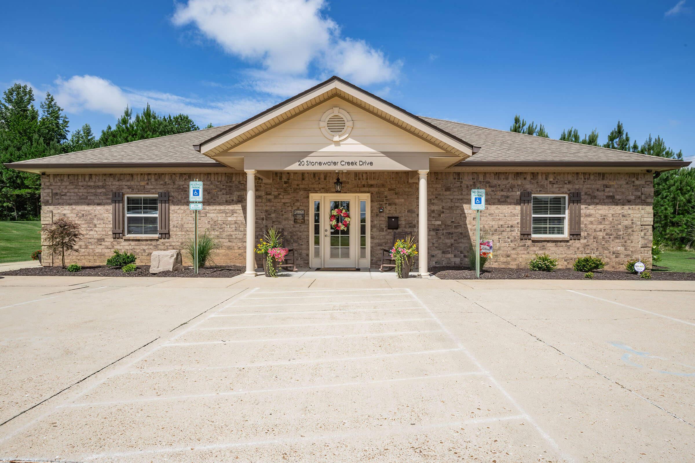 Front view of a brick building with a sloped roof and a welcoming entrance. The facade features decorative landscaping with flower pots, and there are designated parking spaces in front. The sky is bright blue with a few clouds, and a tree line is visible in the background.