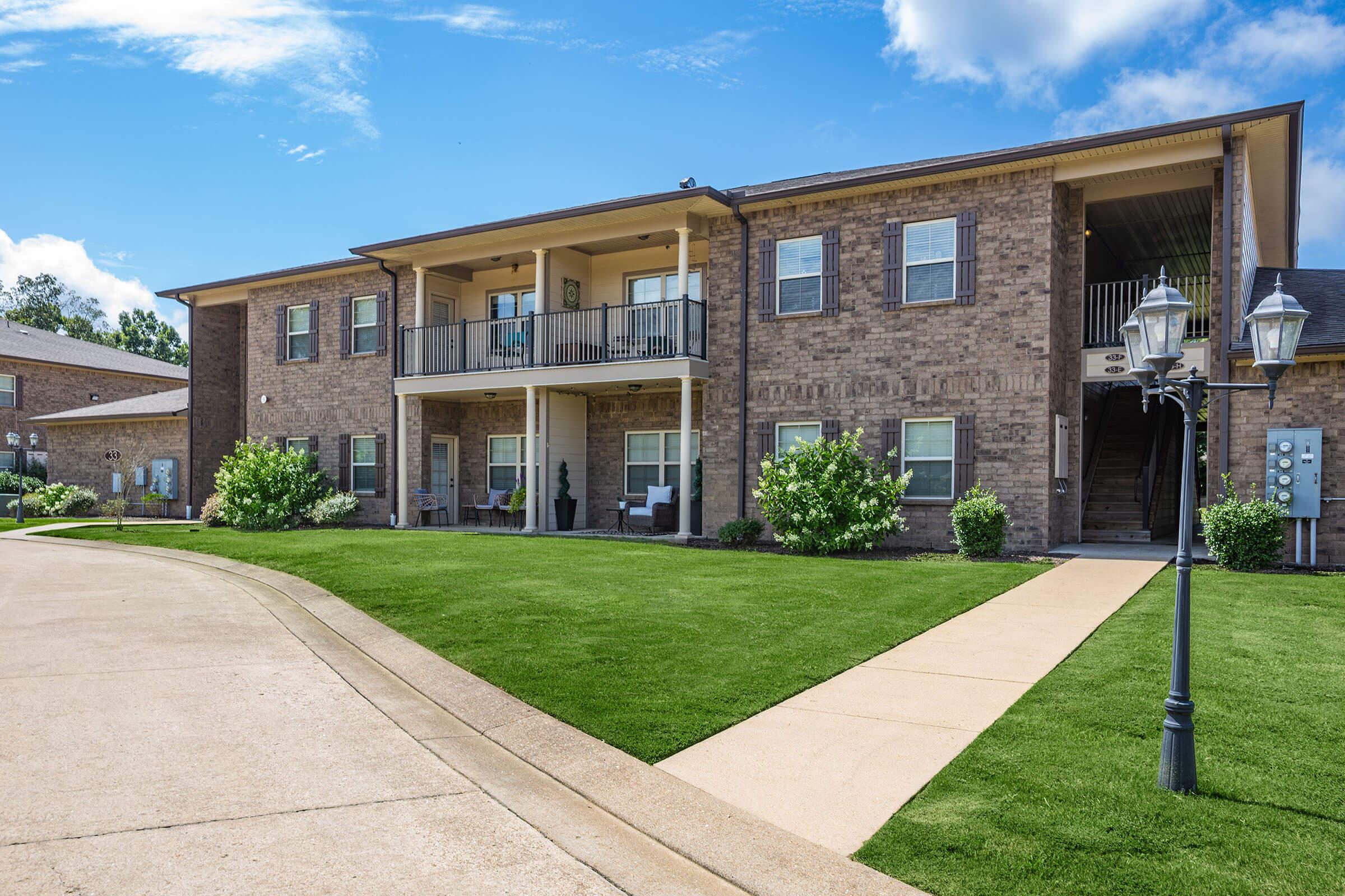 Two-story brick apartment building with balconies, surrounded by neatly manicured green lawns and landscaping. A concrete pathway leads to the entrance, and decorative lampposts are visible along the path. Clear blue sky with a few clouds in the background.