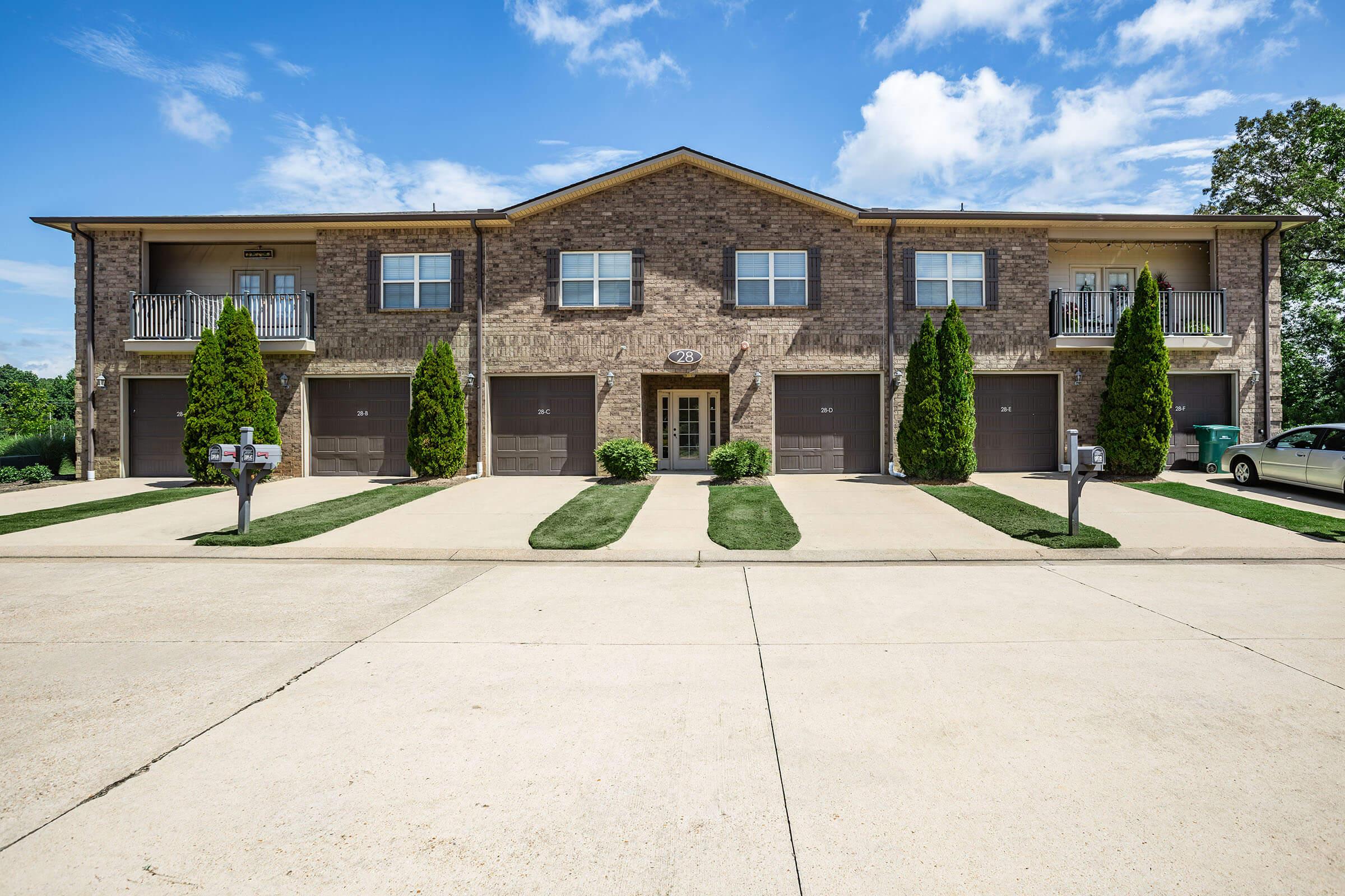 A brick apartment building with two levels, featuring several garages and balconies. The front yard is landscaped with neatly trimmed bushes. A clear blue sky with scattered clouds is visible above. The parking area shows other vehicles and mailboxes along the pathway.