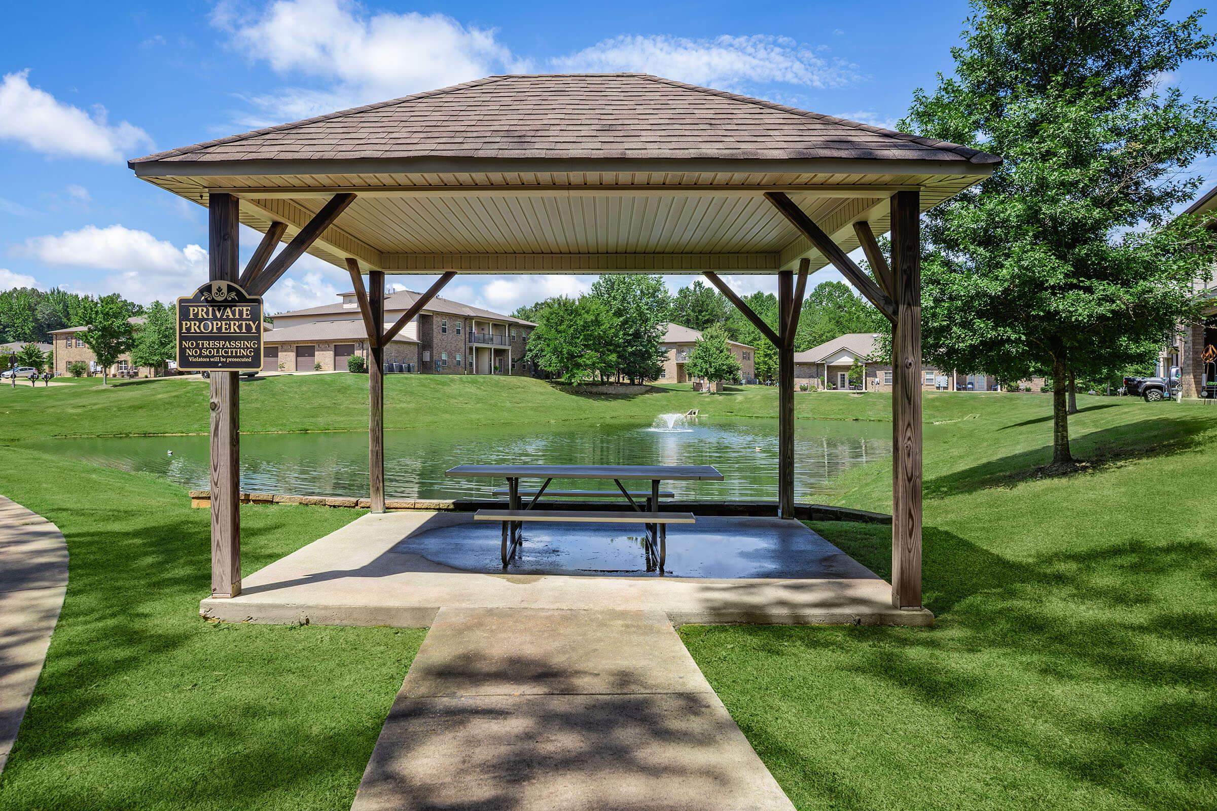 A gazebo with a picnic table is situated near a pond, surrounded by well-maintained grass and trees. A "Private Property" sign is visible on one side of the structure. In the background, there are residential buildings. The sky is partly cloudy, creating a serene outdoor atmosphere.