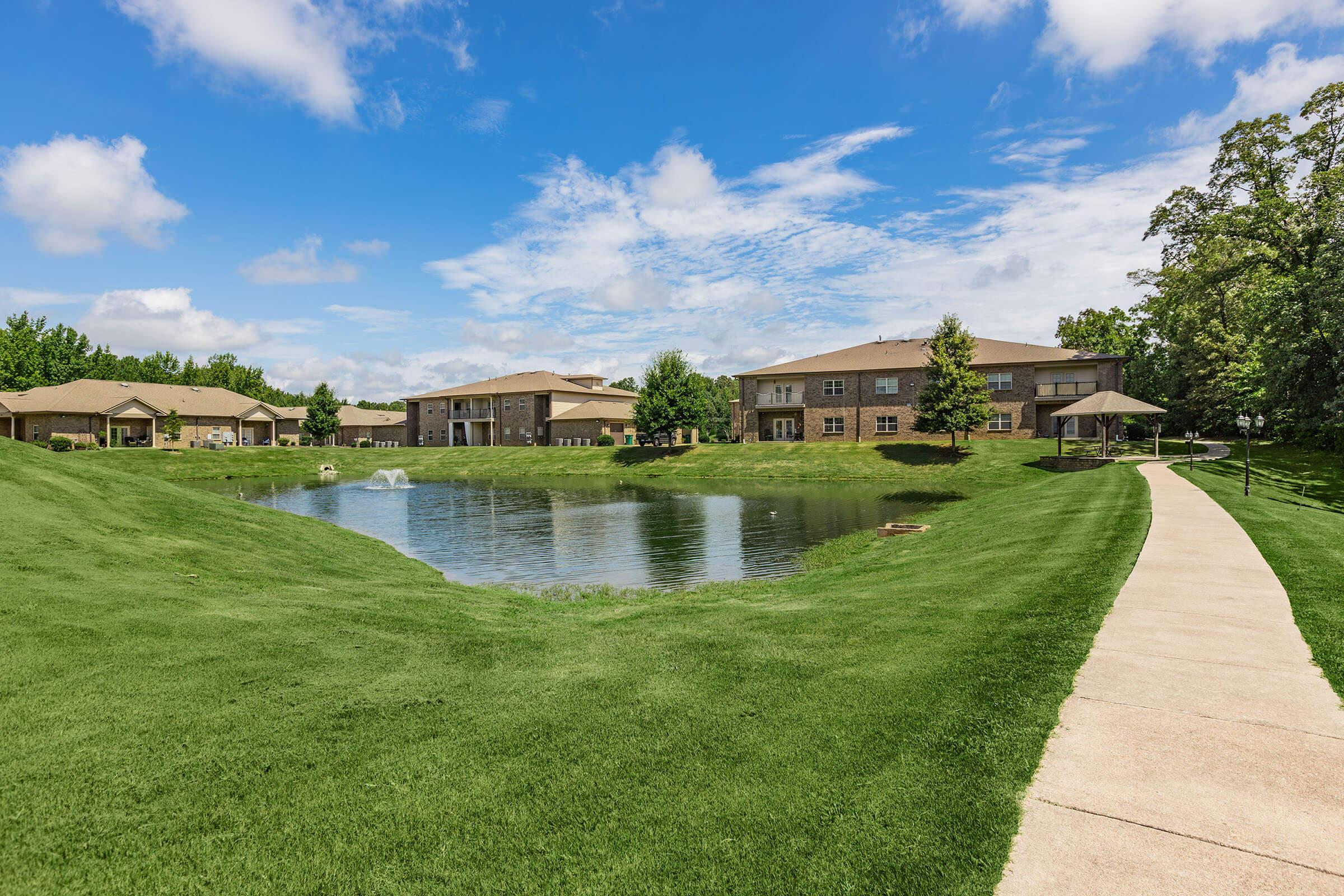A serene landscape featuring a pond surrounded by lush green grass. In the background, there are several residential buildings with a clear blue sky and scattered clouds. A paved walkway runs alongside the pond, inviting leisurely strolls in a peaceful environment.