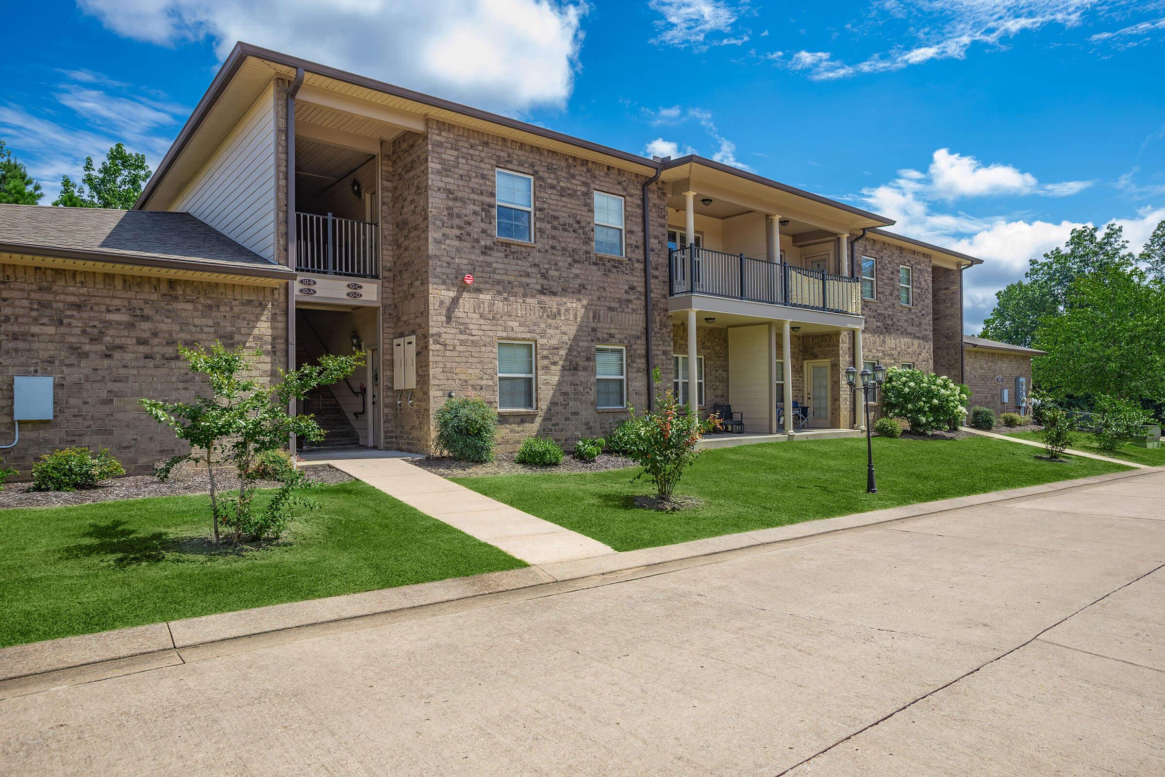 A two-story brick apartment building with a well-maintained lawn, lined with small shrubs and trees. The entrance features a pathway leading to the front doors, and there are balconies on the upper level. The sky is partly cloudy, creating a bright and welcoming atmosphere.