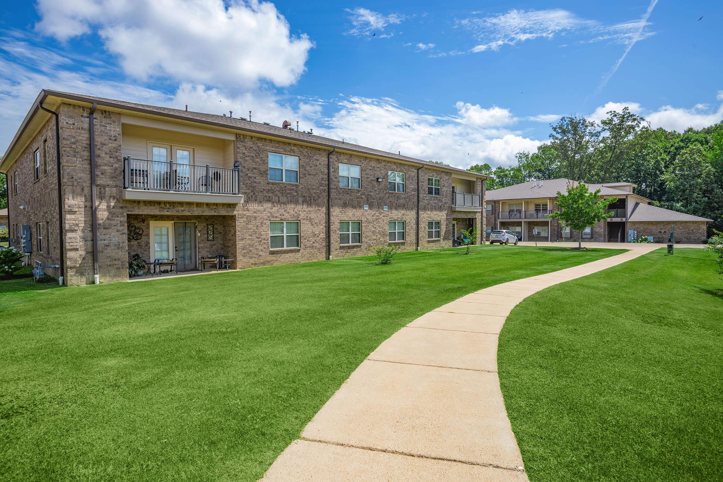 A view of a residential apartment complex surrounded by green grass, featuring two buildings with multiple windows and balconies. A paved walkway leads through the lawn, connecting the buildings under a bright blue sky with scattered clouds.