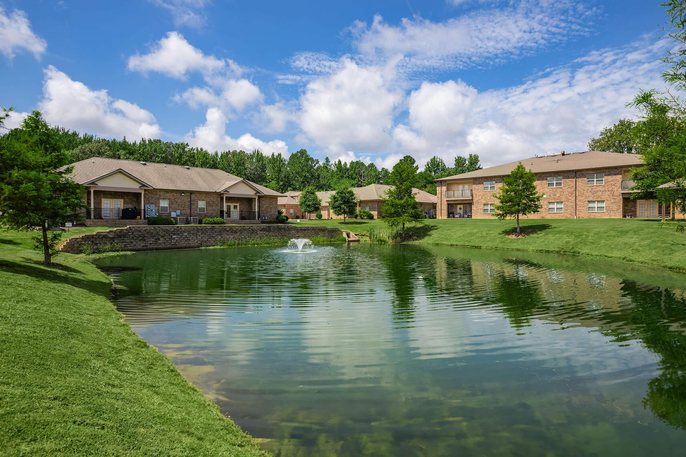 A serene residential area featuring landscaped lawns, a calm pond with a fountain, and apartment buildings in the background under a bright blue sky with fluffy white clouds. Green trees line the edges, creating a peaceful, inviting environment.