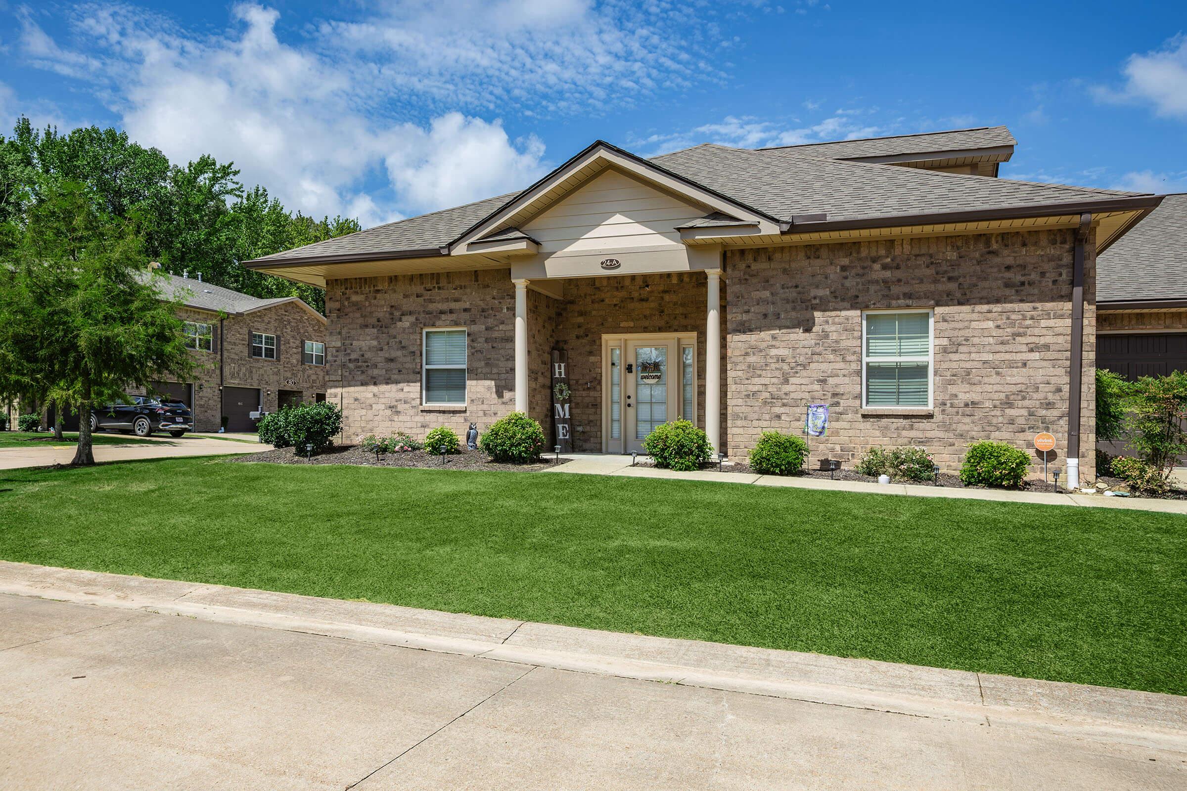 A single-story brick house with a peaked roof and a welcoming front porch. The entrance has a sign that reads "HOME." The lawn features lush, green grass and neatly trimmed bushes. Clear blue skies and fluffy white clouds create a bright, sunny atmosphere in the background.