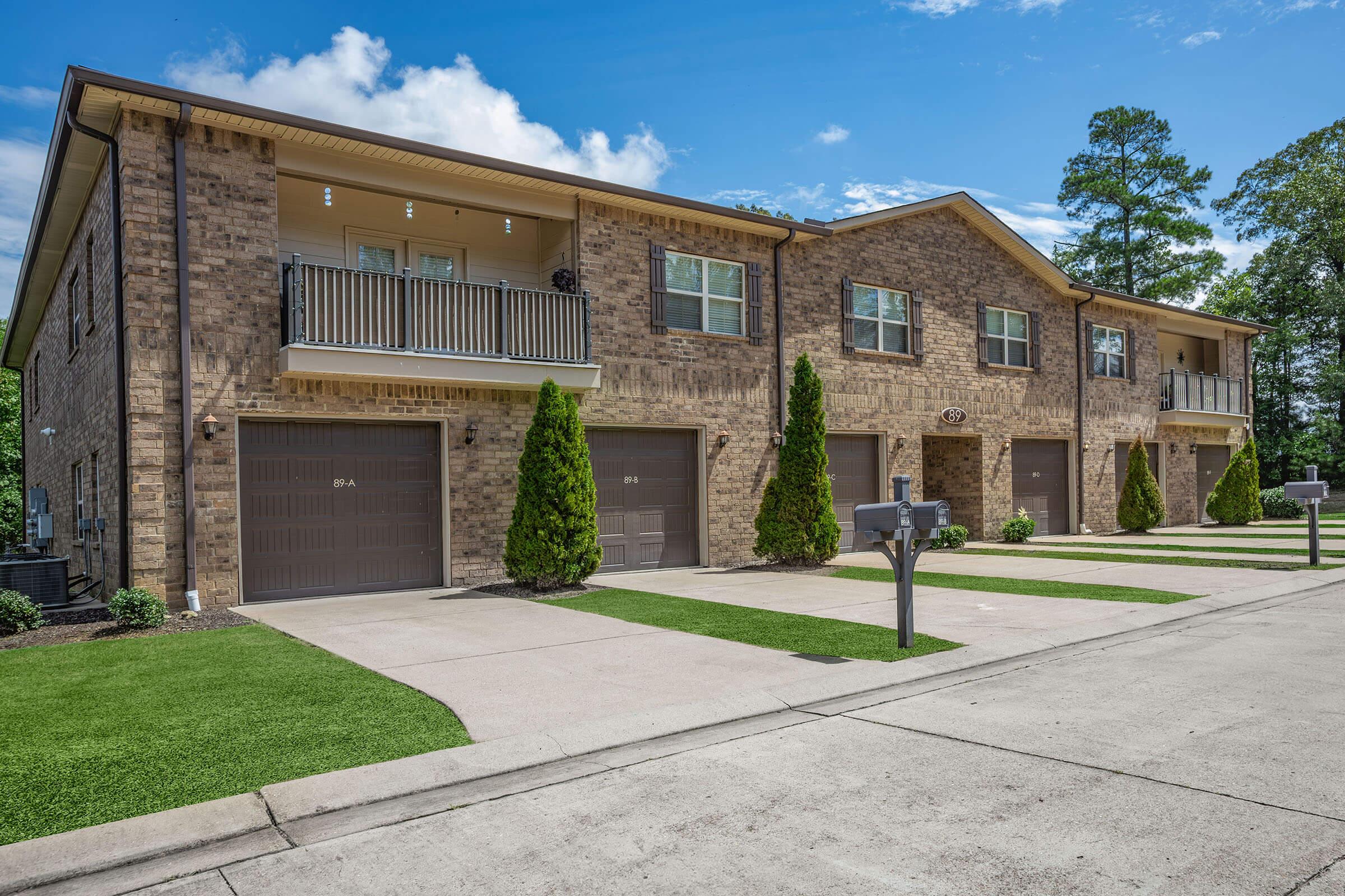 A two-story residential building featuring multiple brick apartments. Each unit has a garage, and the front lawns are neatly maintained with green grass and small shrubs. The driveway is paved, and the sky is clear with scattered clouds in the background.