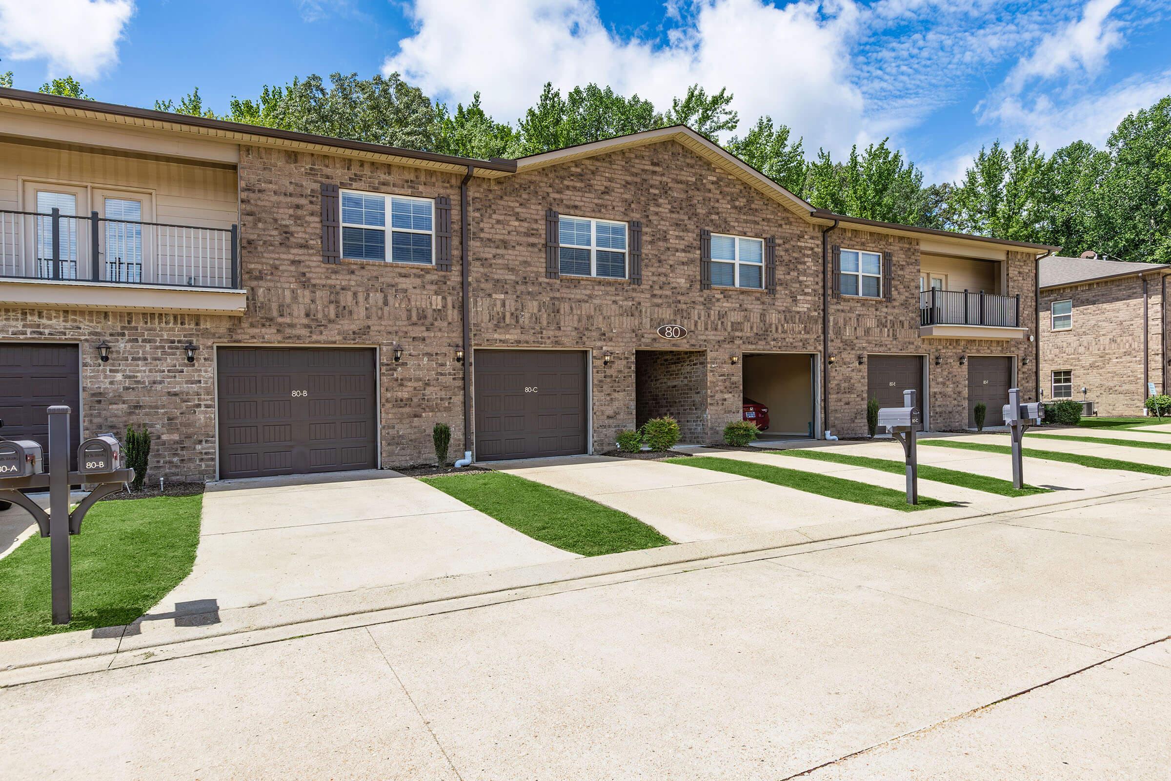 Row of identical brick townhouses with garages, green lawns, and mailboxes in front. The buildings feature second-floor balconies and multiple windows, set against a backdrop of trees and a blue sky with clouds. The driveway is paved and well-maintained.