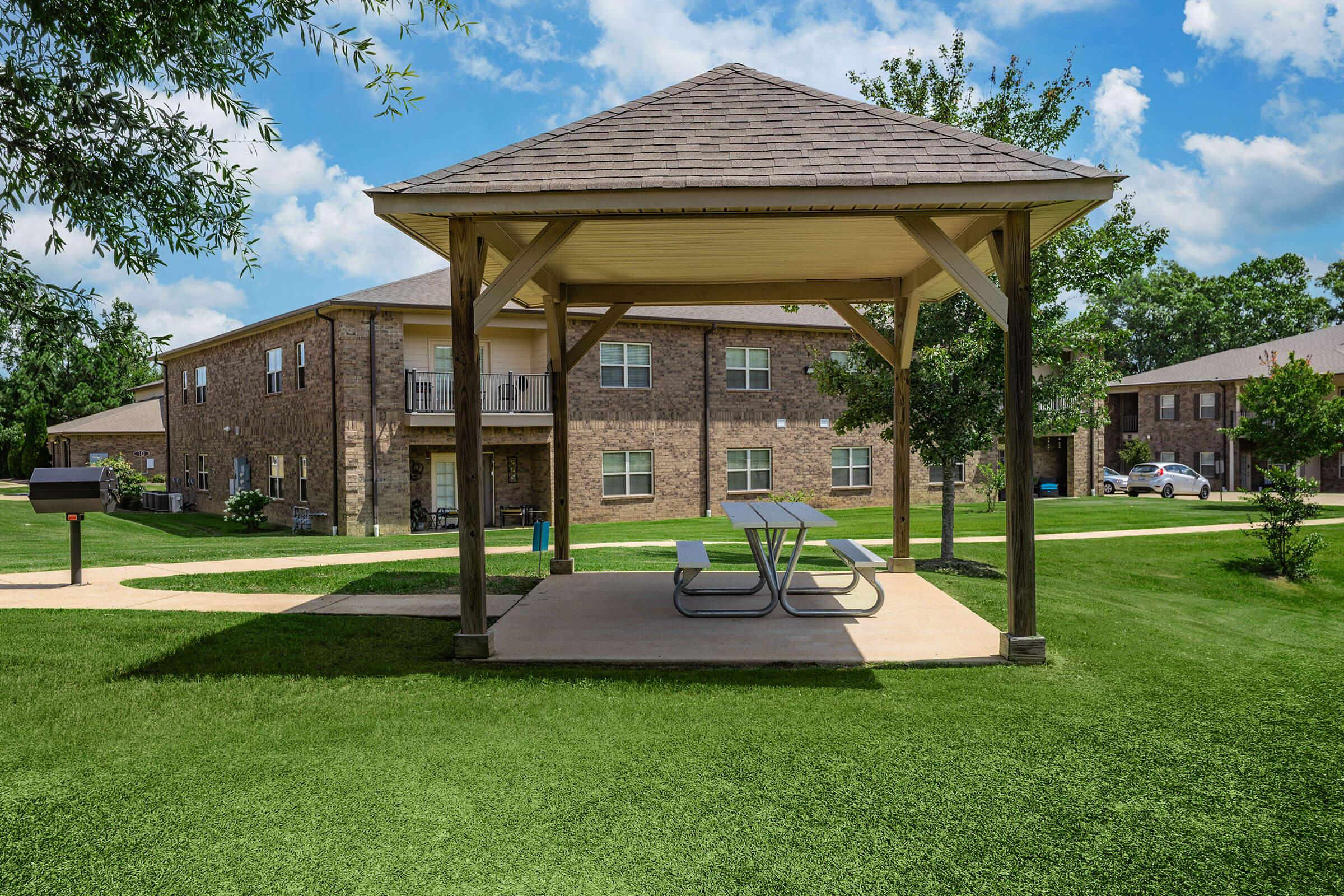 A shaded picnic area with a gazebo featuring two tables, surrounded by well-maintained green grass. In the background, there are multi-story brick apartment buildings under a blue sky with fluffy clouds. The pathway leads to the gazebo and there are trees providing additional greenery.