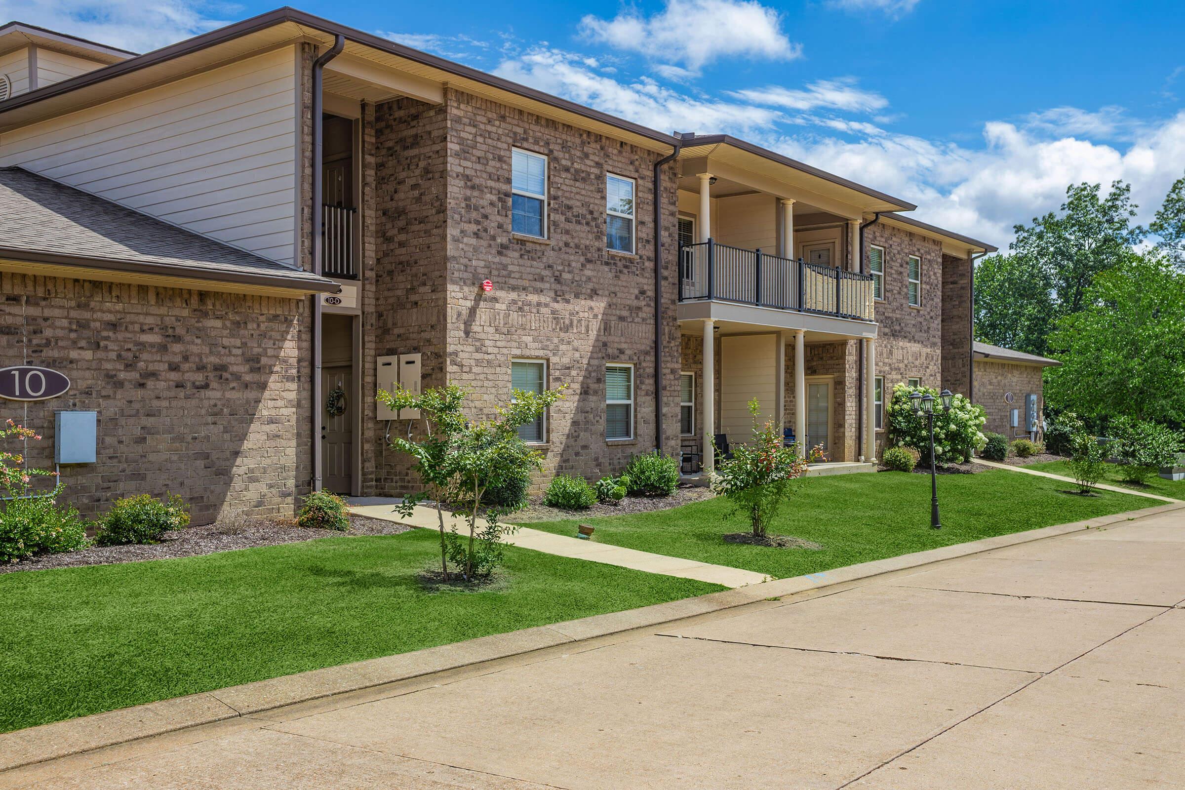 A view of a residential apartment building featuring brick exterior, balconies, and landscaped greenery. The pathway leads to the entrance, with trees and shrubs enhancing the exterior aesthetics under a partly cloudy blue sky. 
