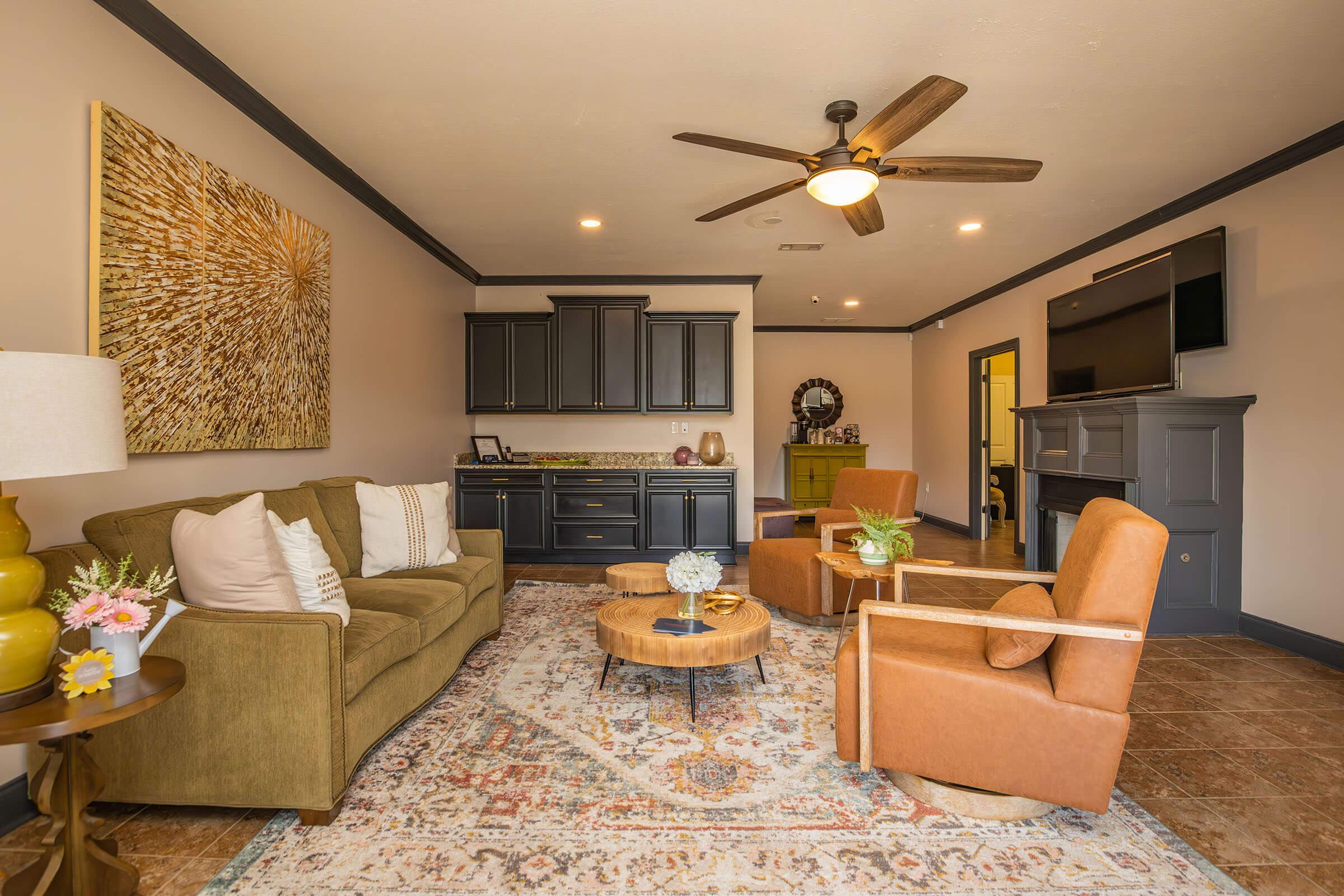 A cozy living room featuring a green sofa and two brown armchairs around a wooden coffee table. There is a decorative area rug, a modern ceiling fan, and a kitchenette in the background. The walls are a soft beige with dark trim, and a large piece of artwork hangs above the sofa.