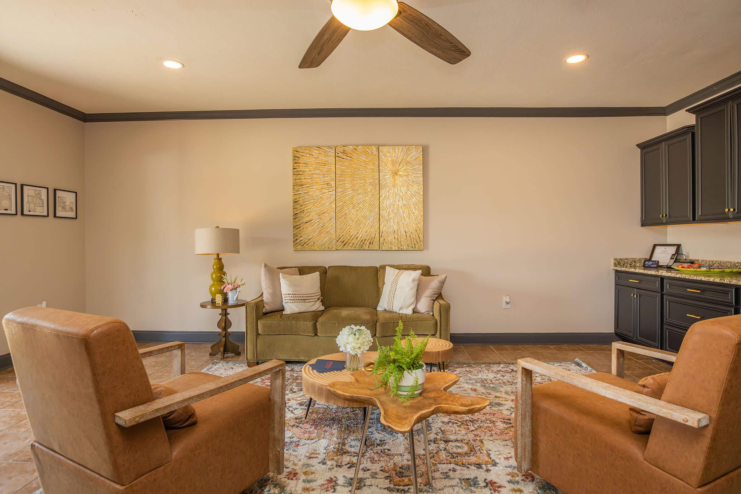 A welcoming living room featuring a green sofa with pillows, two brown armchairs, and a wooden coffee table. A ceiling fan is overhead, and there’s an abstract gold artwork on the wall. The space is decorated with plants and warm, neutral tones, creating a cozy atmosphere.