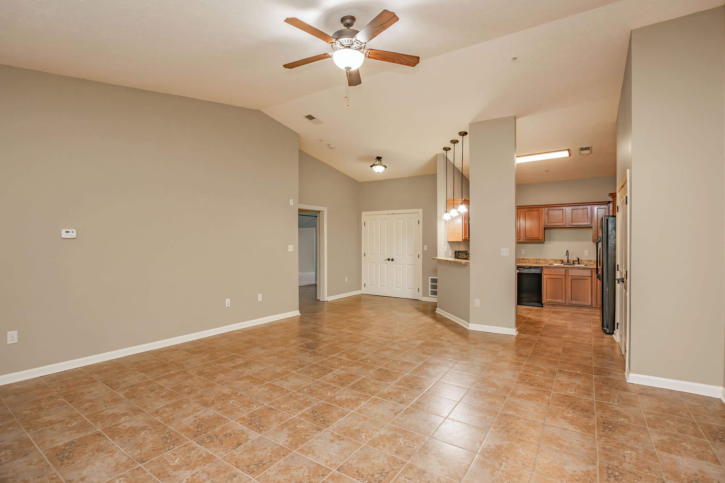 Spacious living area with high ceilings, a ceiling fan, and tiled flooring. Features an open layout connecting to a modern kitchen with wooden cabinets and stainless steel appliances. Natural light enters through large windows, creating a bright atmosphere. The walls are painted in neutral tones.