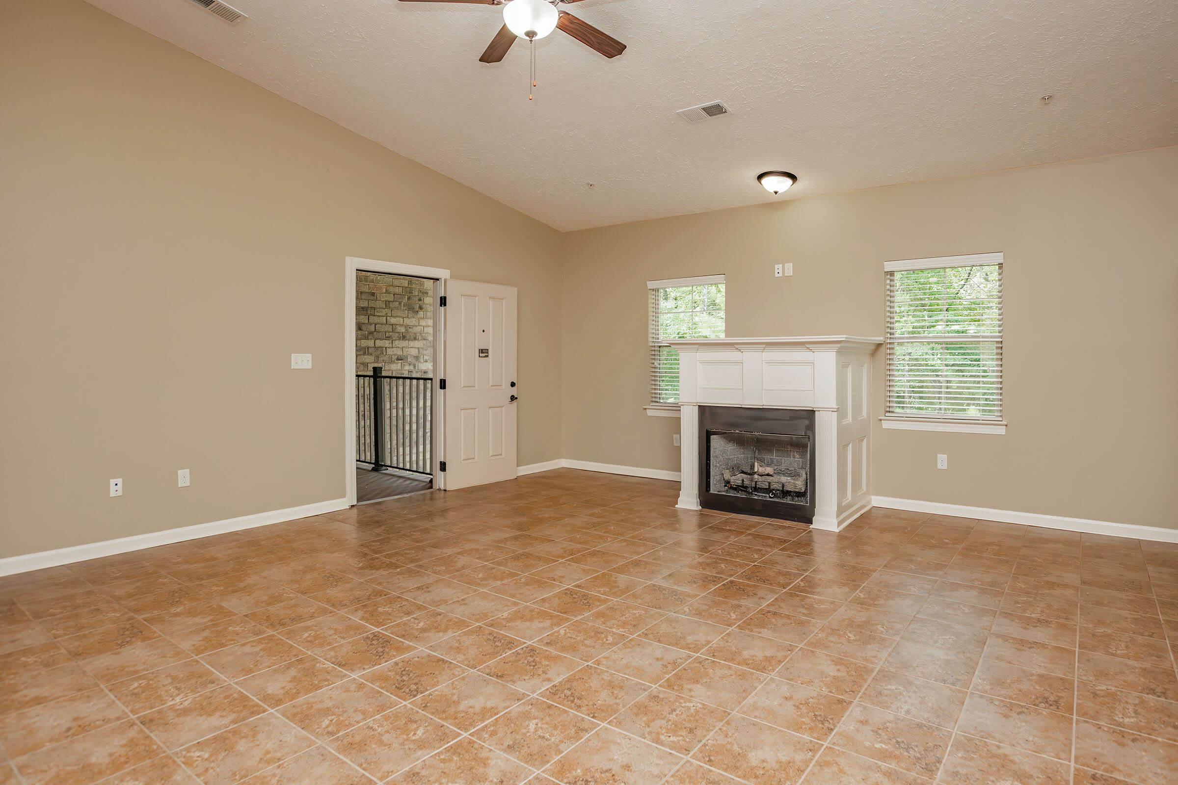 Spacious living room with tiled flooring, a ceiling fan, and a decorative fireplace. Two windows provide natural light, and there is a doorway leading to a balcony area. The walls are painted a light beige color, creating a warm and inviting atmosphere.