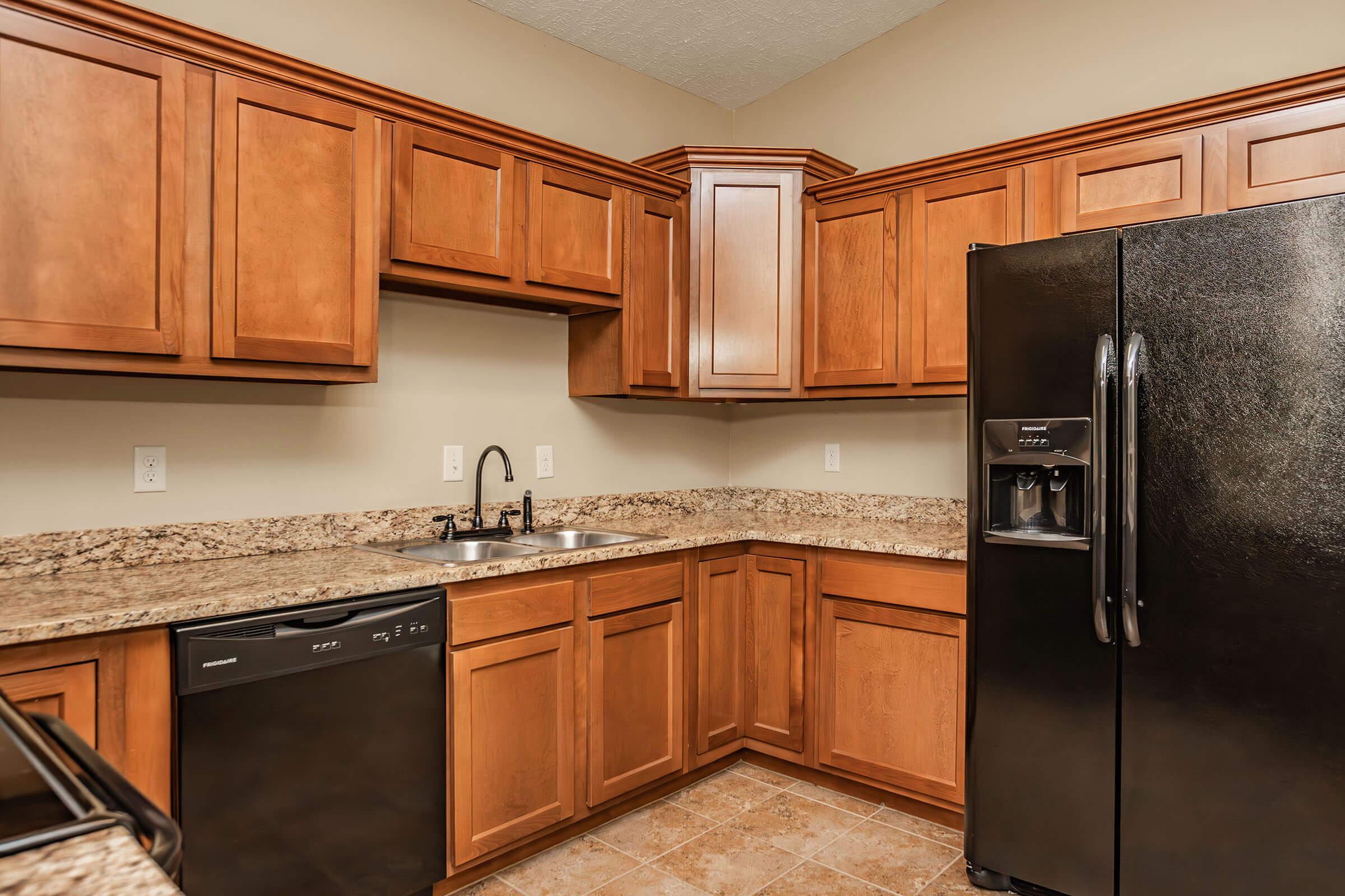 A modern kitchen featuring wooden cabinetry, a black refrigerator, a sink with a faucet, and a dishwasher. The countertops are made of granite, and the floor is tiled. The kitchen has warm lighting and an open layout, creating a spacious feel.