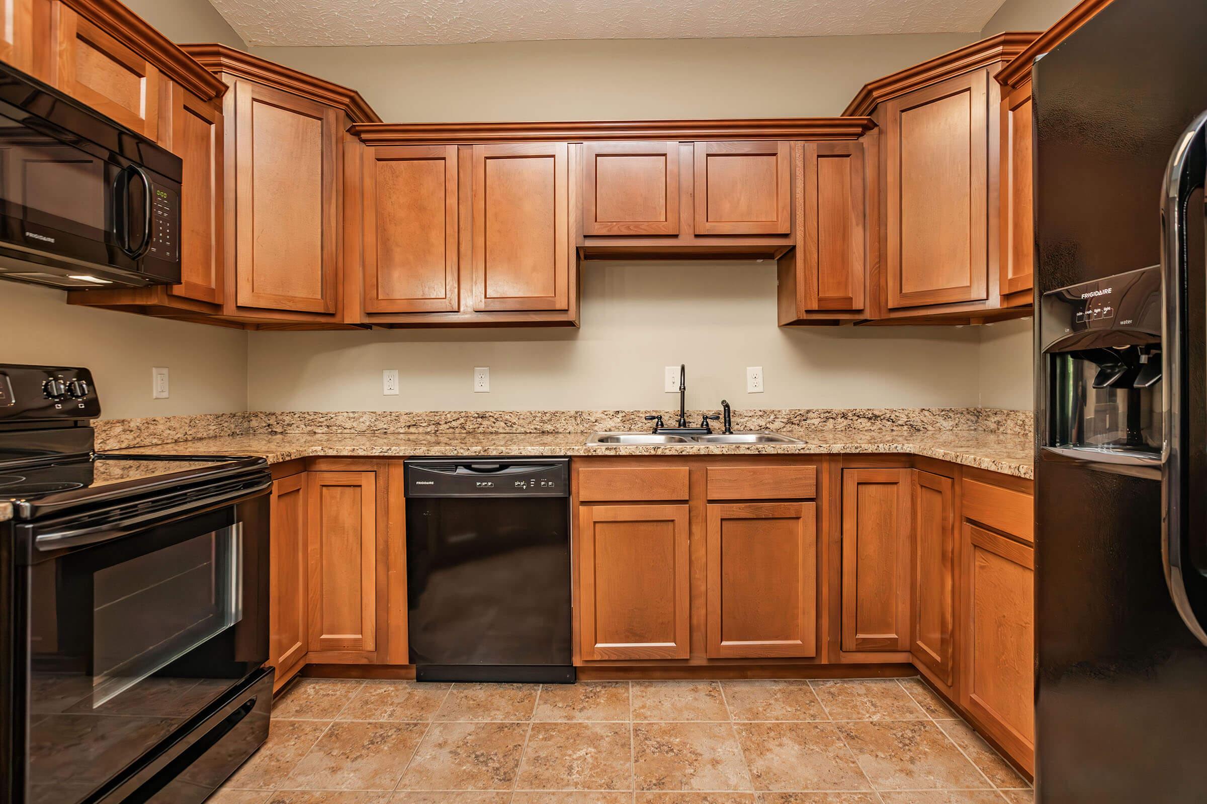 A modern kitchen featuring wooden cabinets, a black dishwasher, and a black stove. The countertops are made of speckled granite, and there’s a double sink with a faucet. A black refrigerator is also visible, and the floor is tiled with light-colored tiles. The walls are painted in a neutral tone.