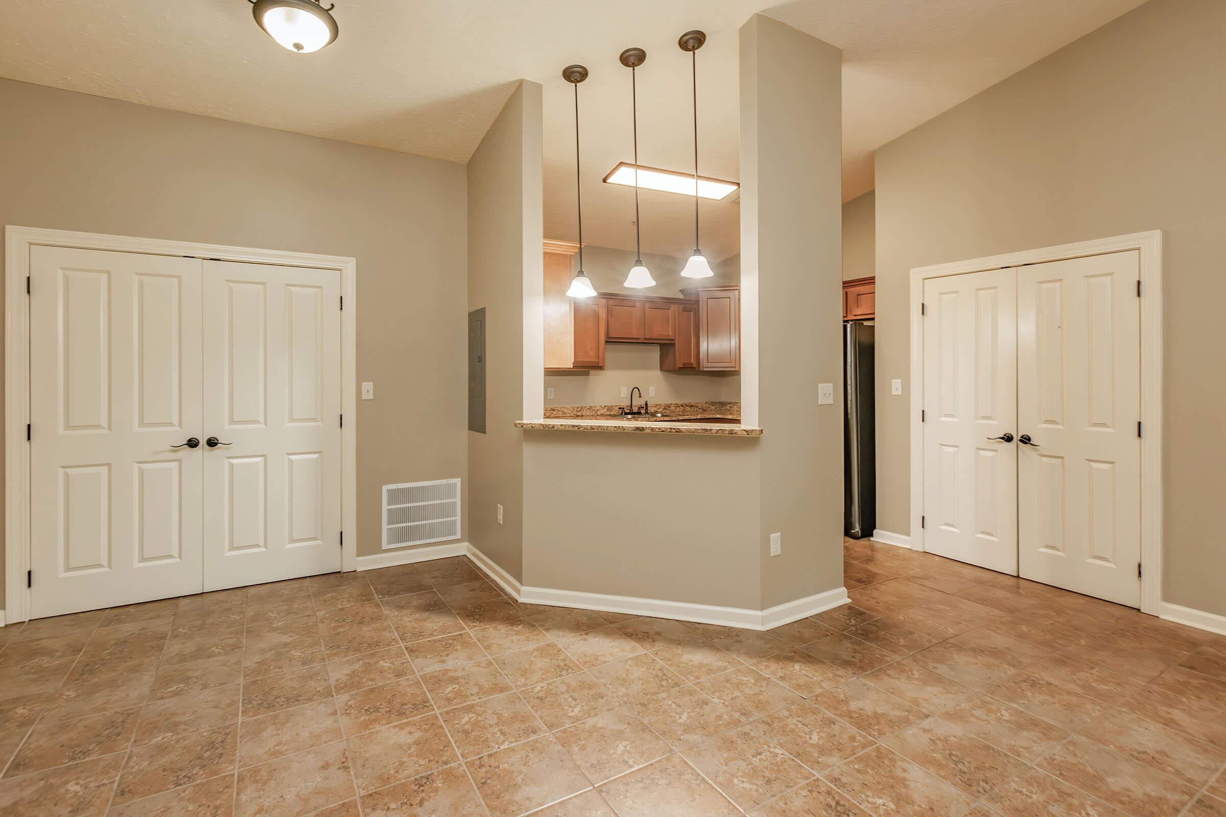 A spacious, well-lit interior featuring beige walls and tile flooring. The kitchen area has a countertop with three pendant lights overhead and wooden cabinets. Two closed doors are visible on the left, and a black appliance sits next to the right door. The layout offers an open, inviting atmosphere.