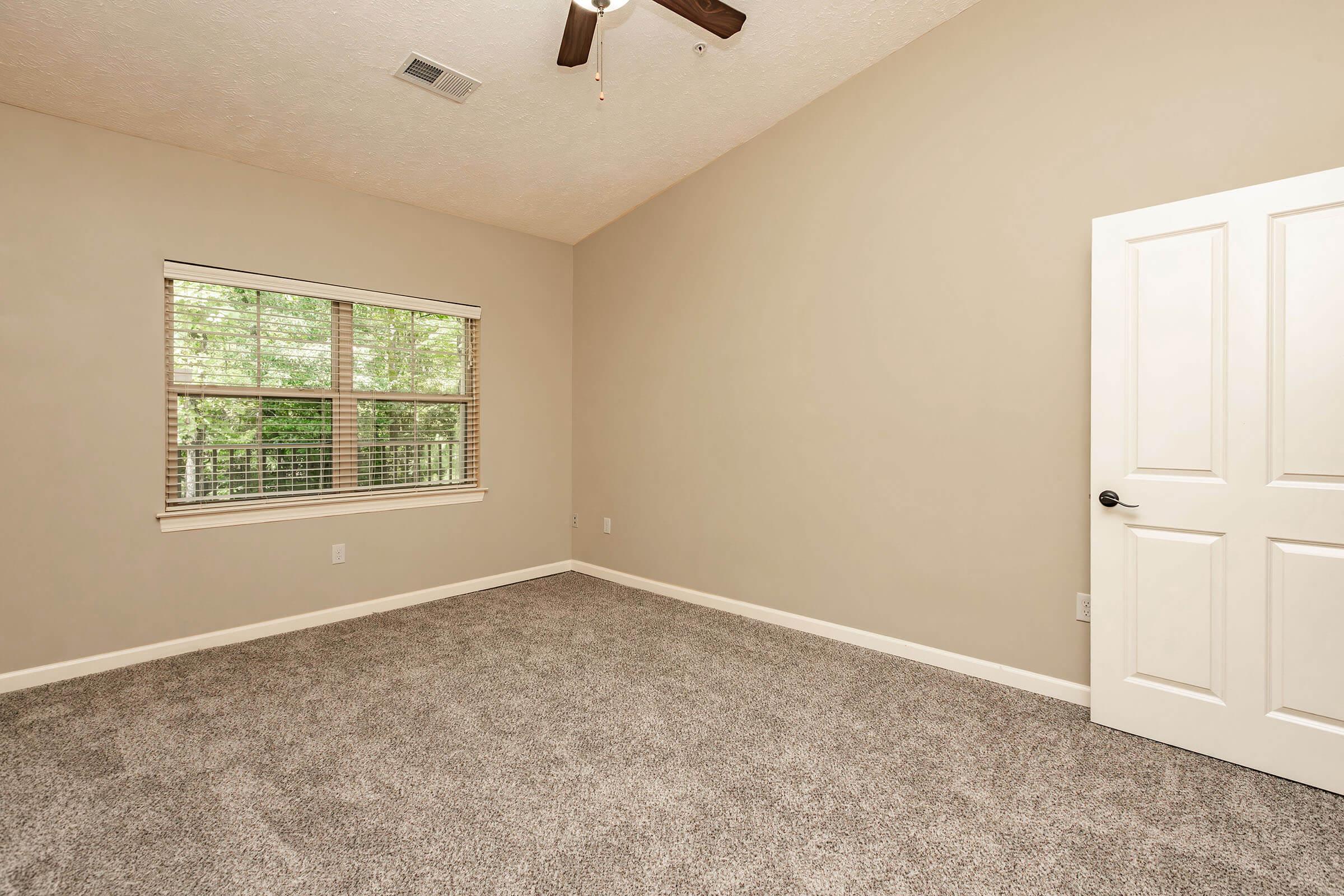 A spacious, empty room with beige walls and a light-colored carpet. It features a large window with blinds allowing natural light to enter, and a ceiling fan. The room also has a white door leading to another space, contributing to a modern and clean aesthetic.