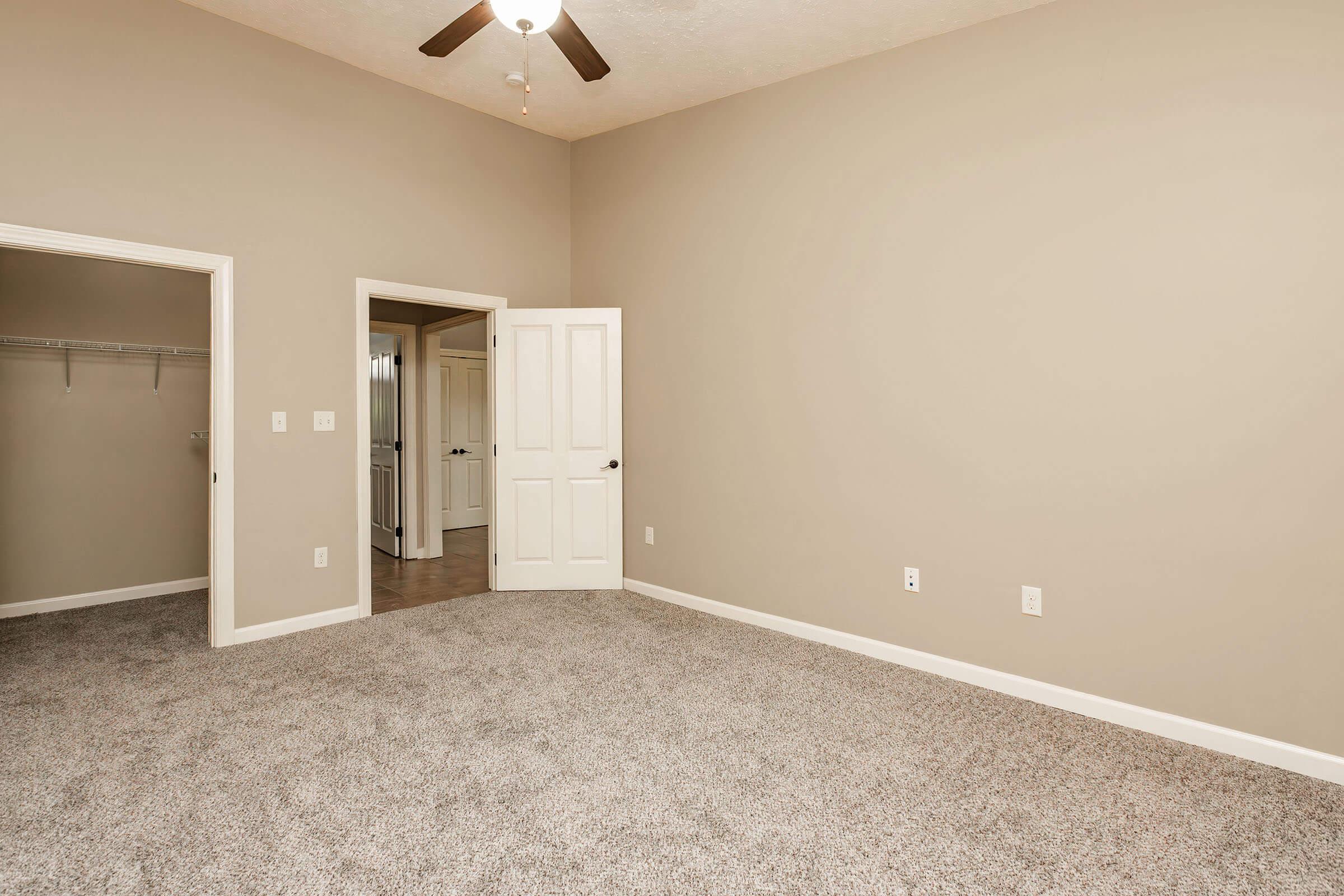 A spacious, unfurnished room featuring light beige walls and carpeted flooring. It includes a ceiling fan and an open doorway leading to a closet on one side, while another door opens to a hallway. The overall atmosphere is bright and neutral, suitable for various decorating styles.