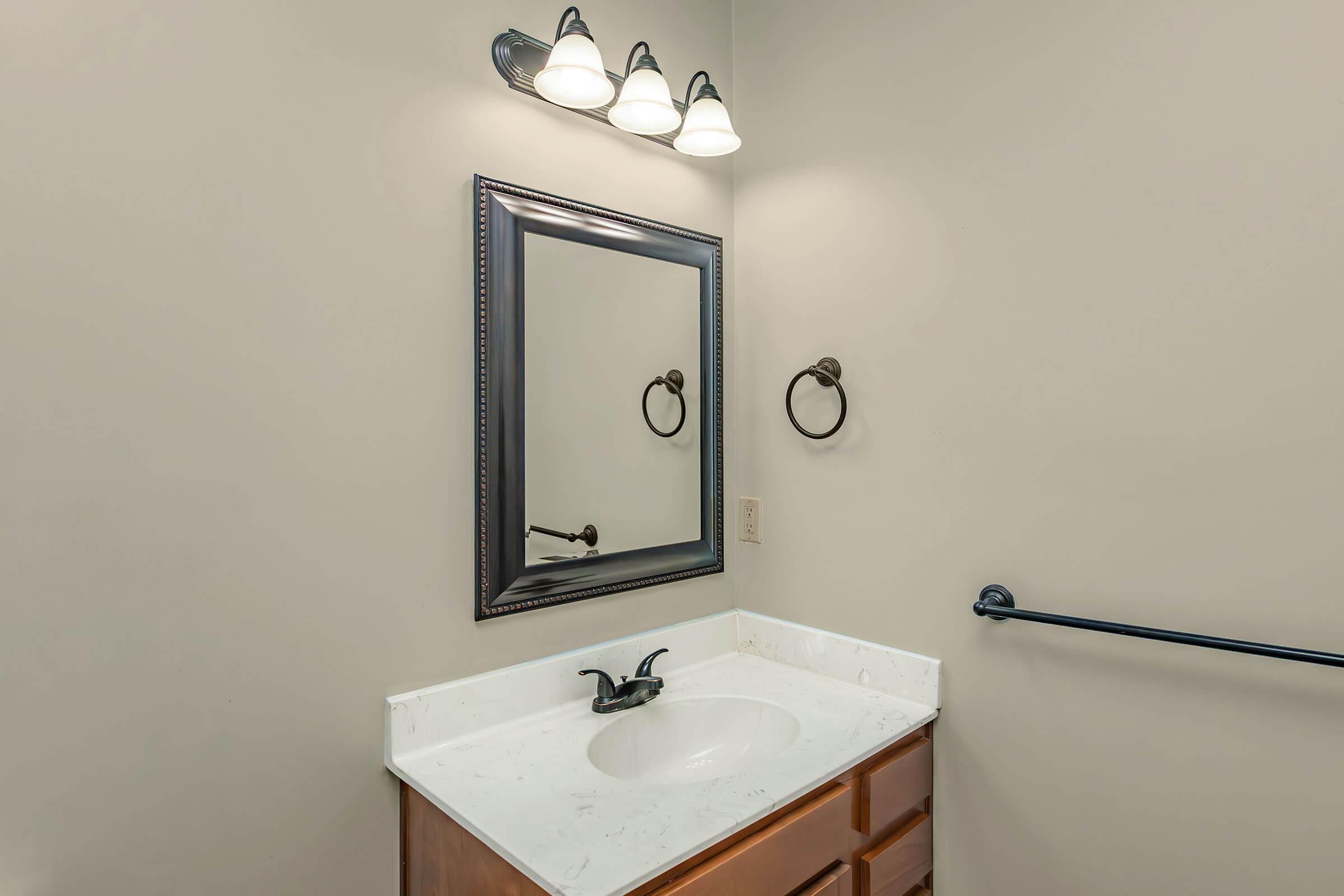 A bathroom with a light-colored wall, featuring a wooden vanity with a sink, a large mirror framed in black, and three light fixtures above. There's a decorative towel ring on the wall and a grab bar beside the sink. The overall ambiance is clean and simple.