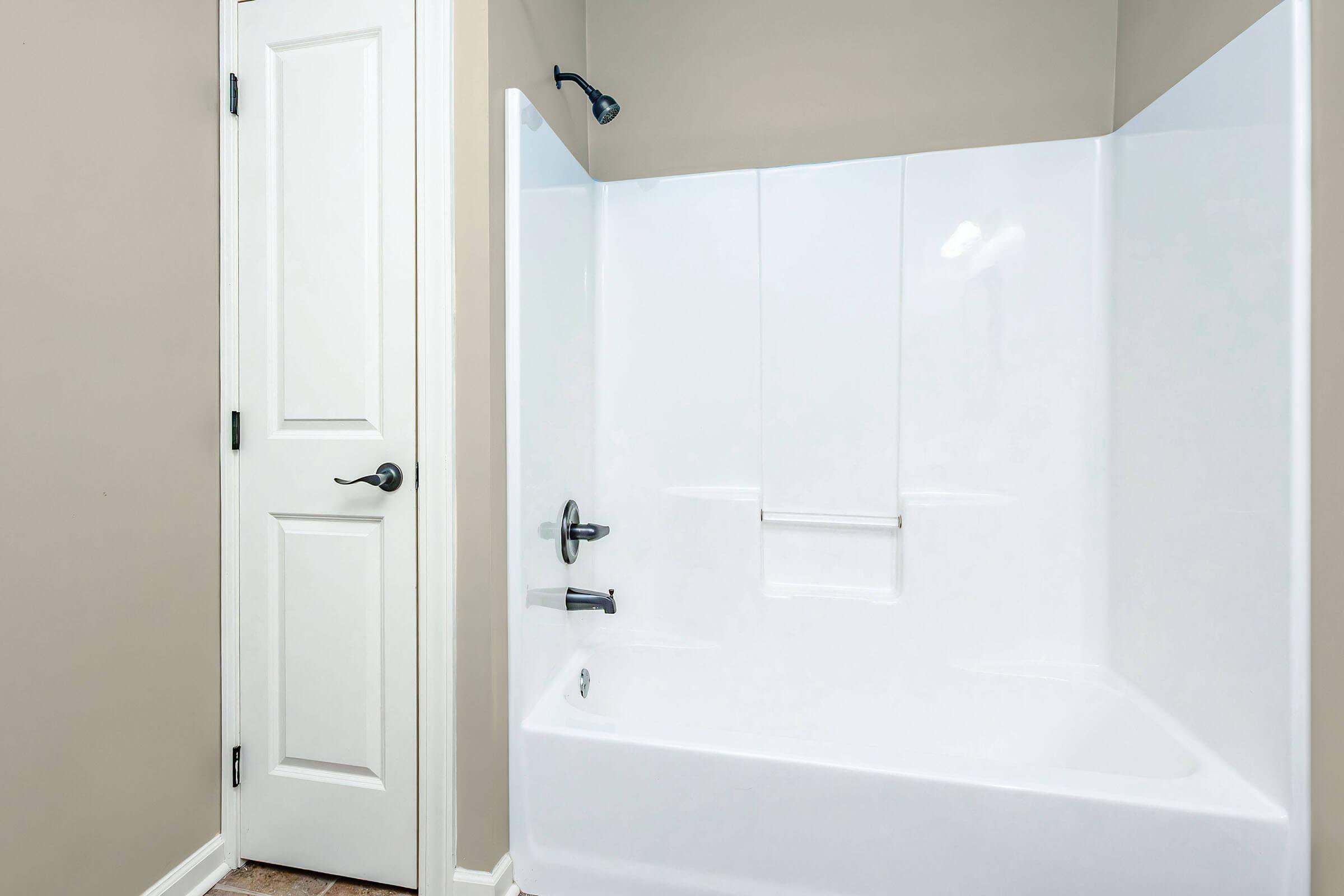 A clean, uncluttered bathroom featuring a white bathtub and shower enclosure. The walls are painted a neutral color, and there is a closed wooden door on the left. A showerhead is mounted on the wall, and a small shelf is visible on the side of the tub. The overall atmosphere is fresh and inviting.
