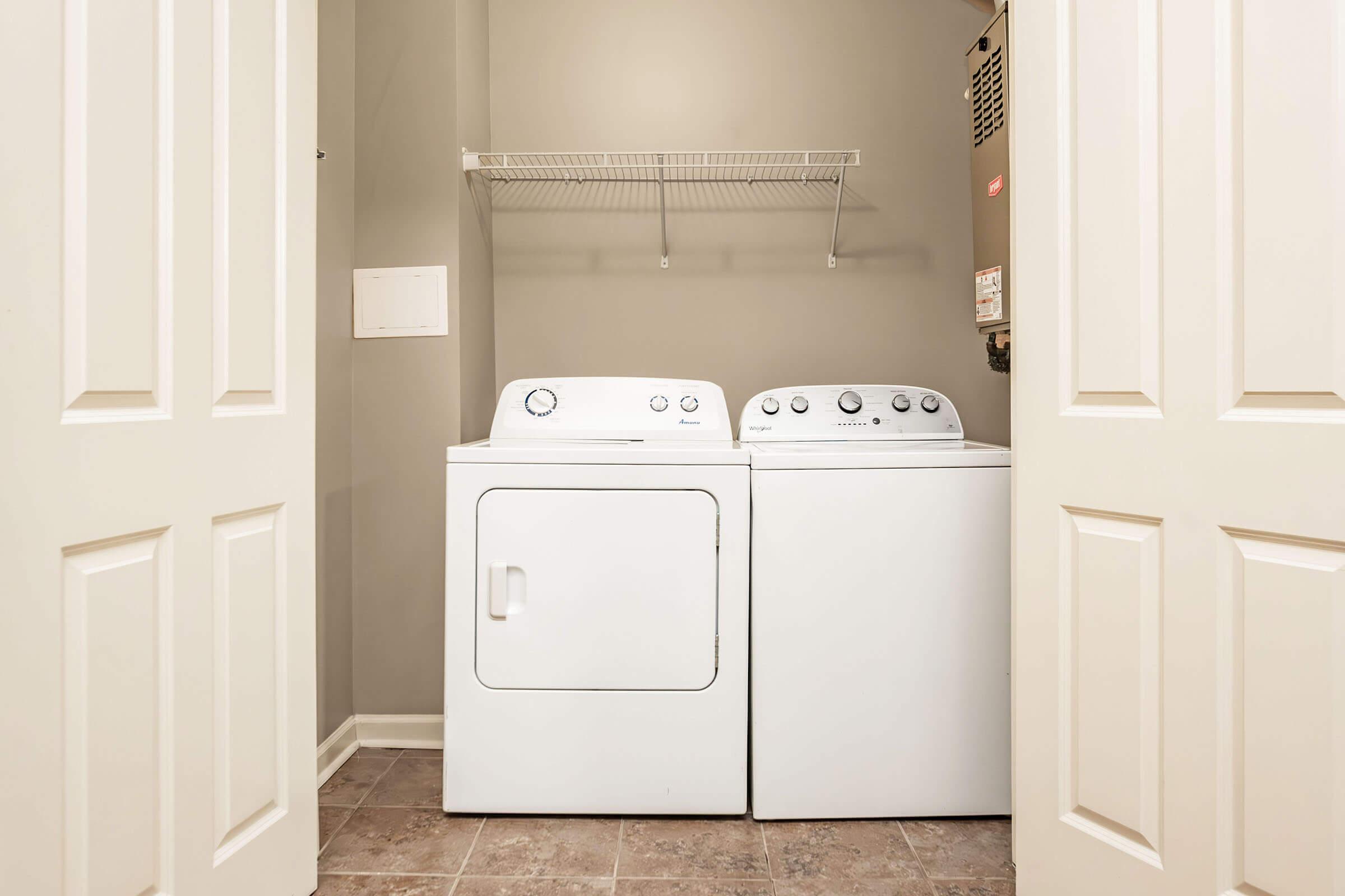 A laundry room featuring a stackable washer and dryer set side by side on a tiled floor. Light-colored walls and a shelf above the appliances provide storage space. The room is accessed through double doors, creating a clean and functional space for laundry tasks.